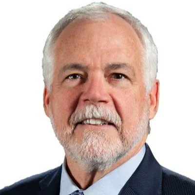 Close-up of an older man with white hair and a beard, wearing a dark suit and light blue shirt, smiling softly against a plain white background.