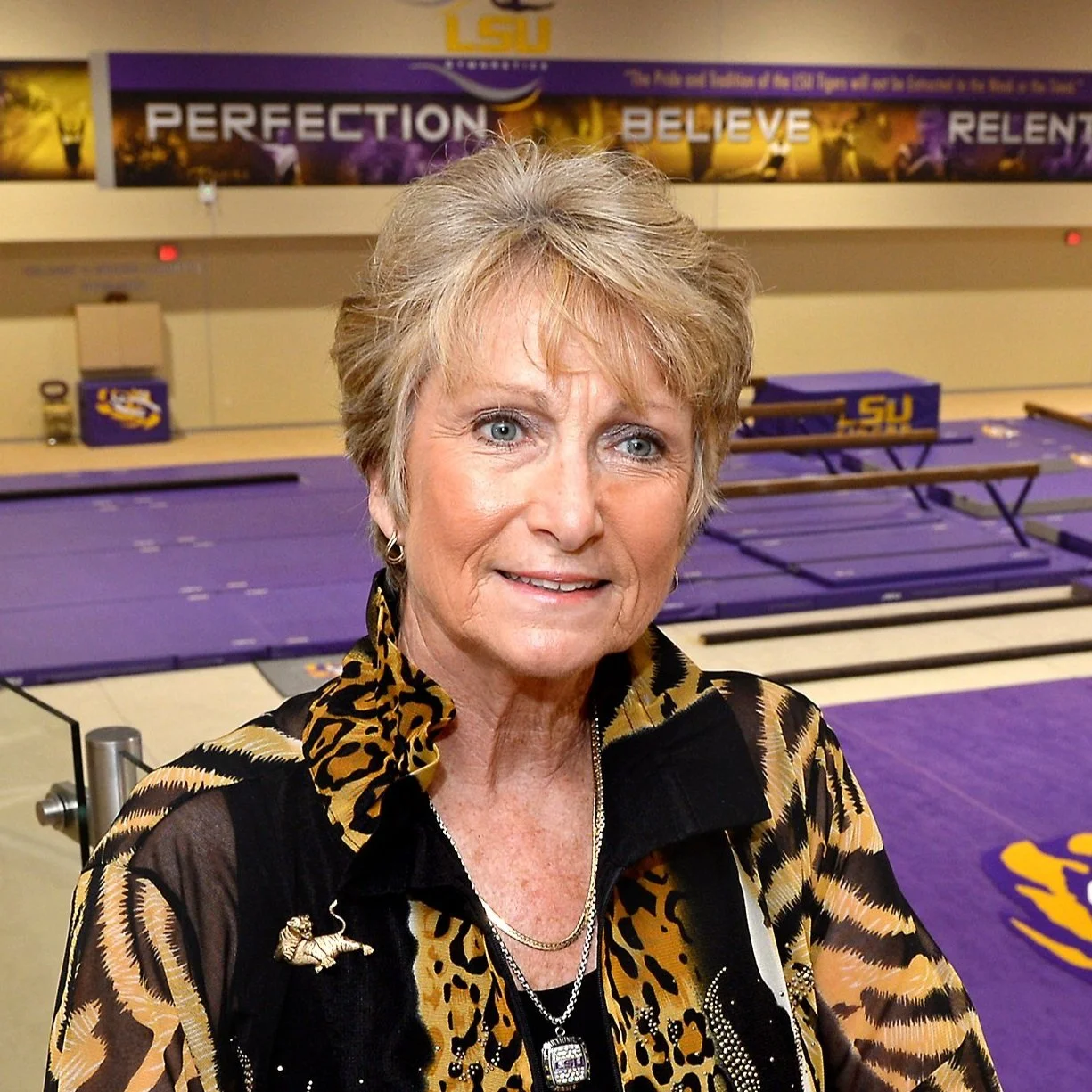 An older woman with short blonde hair, wearing a leopard print jacket, posing inside an athletic facility with purple gymnastics mats and 'LSU' branding in the background.