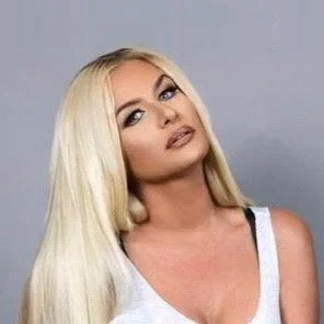 A woman with long blonde hair, makeup, and a white sleeveless top posing for the camera against a plain background.