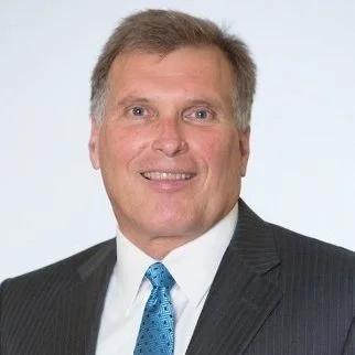 Headshot of a man in a business suit with a blue tie, smiling against a plain background.