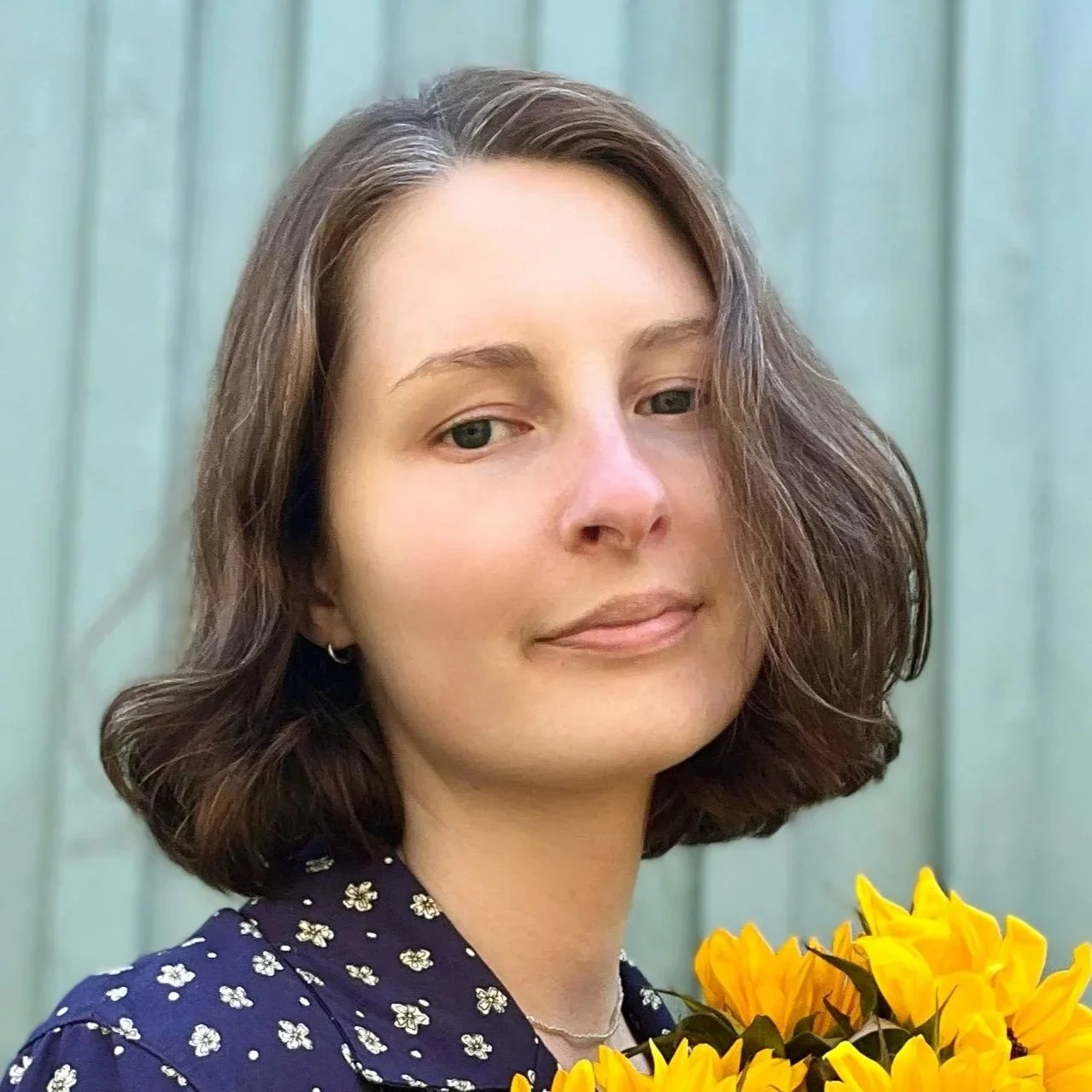 A woman with shoulder-length brown hair, wearing a navy blue blouse with white floral pattern, holding a bouquet of yellow sunflowers, standing in front of a light green wooden background.