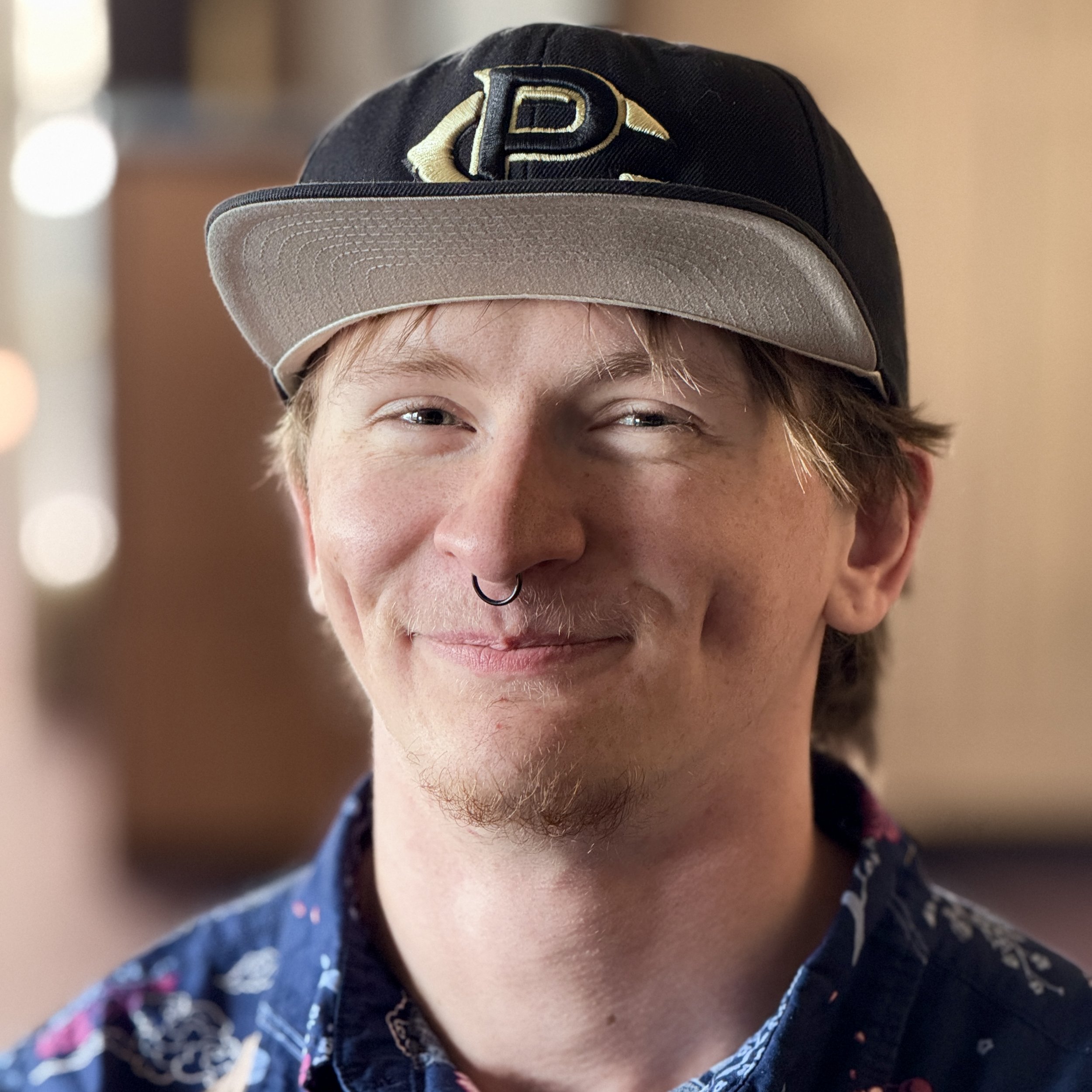 Close-up of a person wearing a black baseball cap with a beige brim and an embroidered letter D in gold outlined in black on the front, smiling.