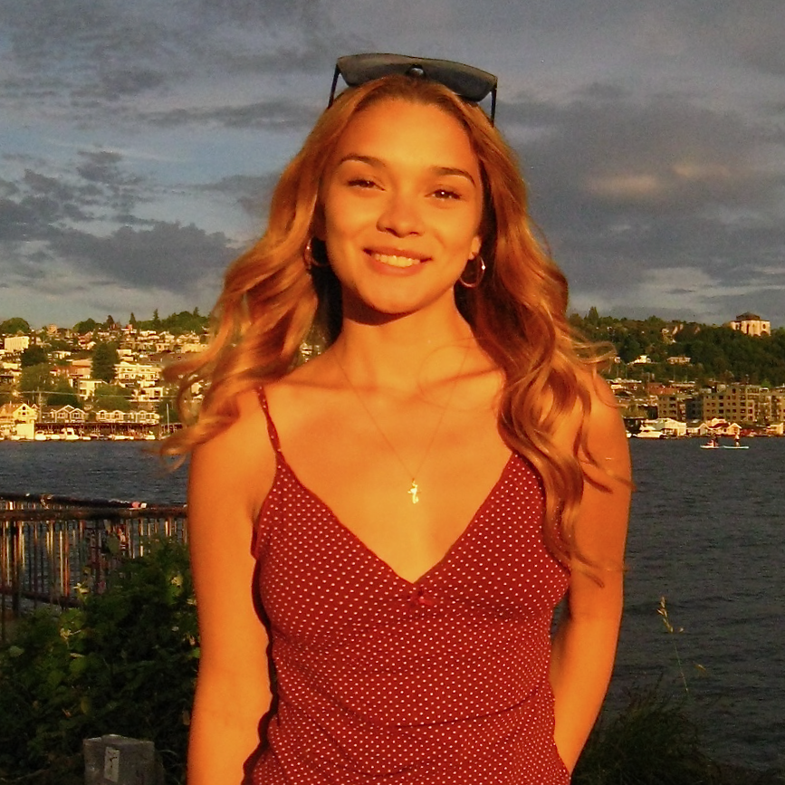 A young woman with long wavy blond hair, wearing a red polka dot sleeveless dress, standing outdoors near a body of water with a cityscape in the background.