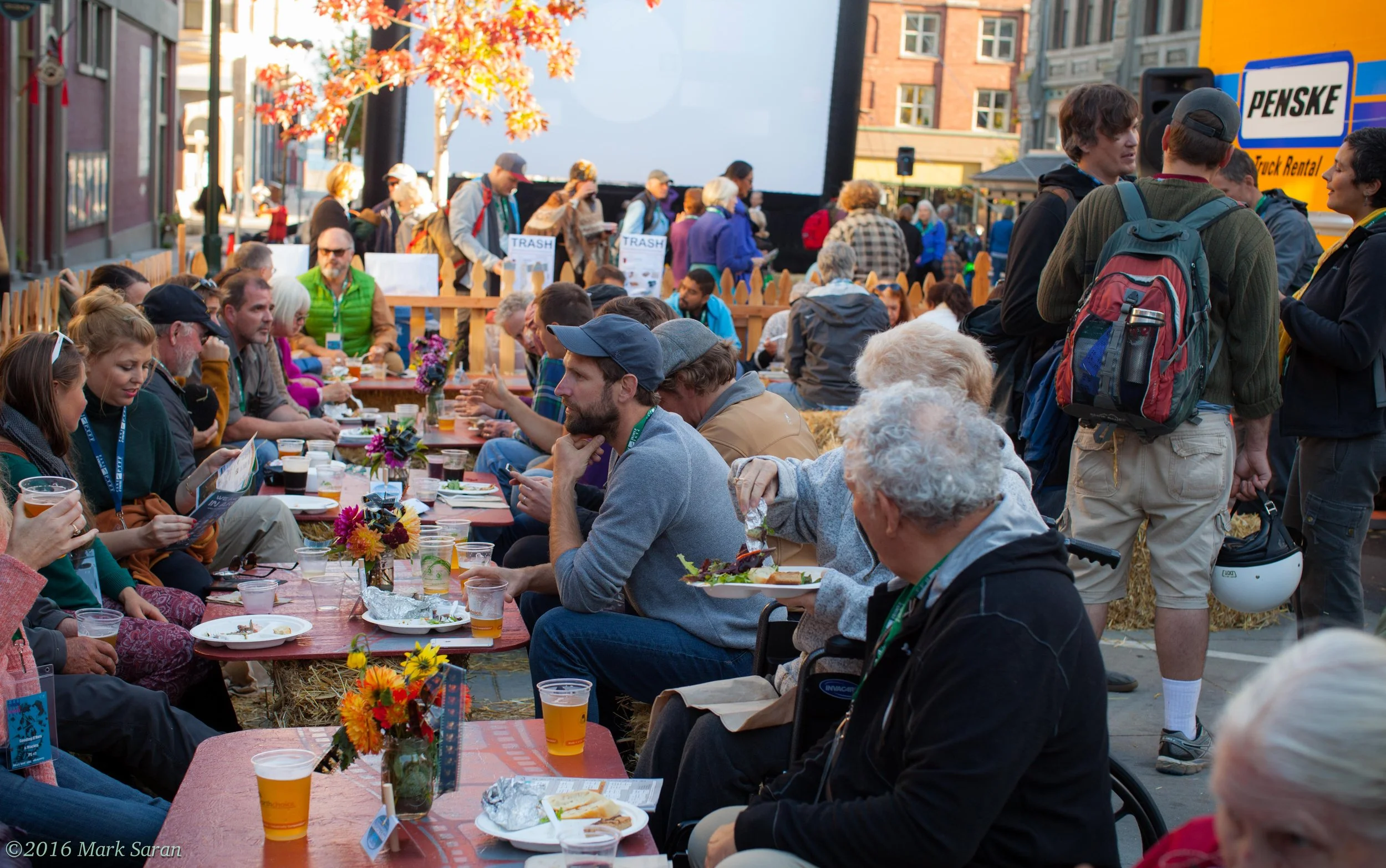 People gathered outdoors at a picnic or festival, seated at a long table with food and drinks, some standing or walking around, with trees, buildings, and a large screen or projector in the background.