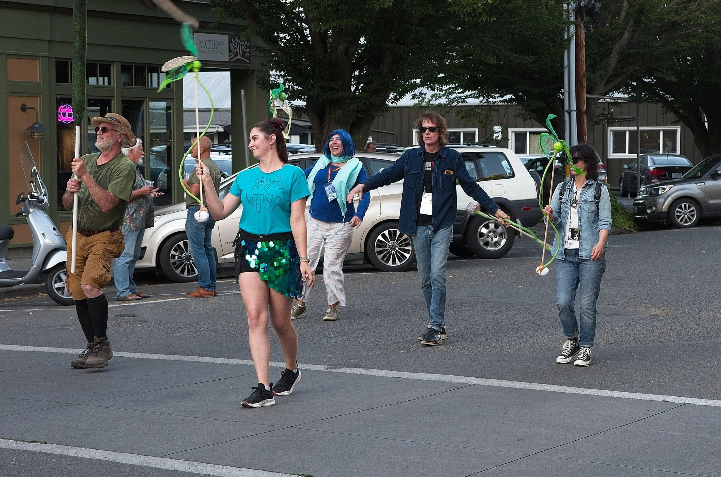 Group of people walking outdoors carrying artificial flowers with googly eyes, smiling and engaging in a playful parade or event, with parked cars and buildings in the background.