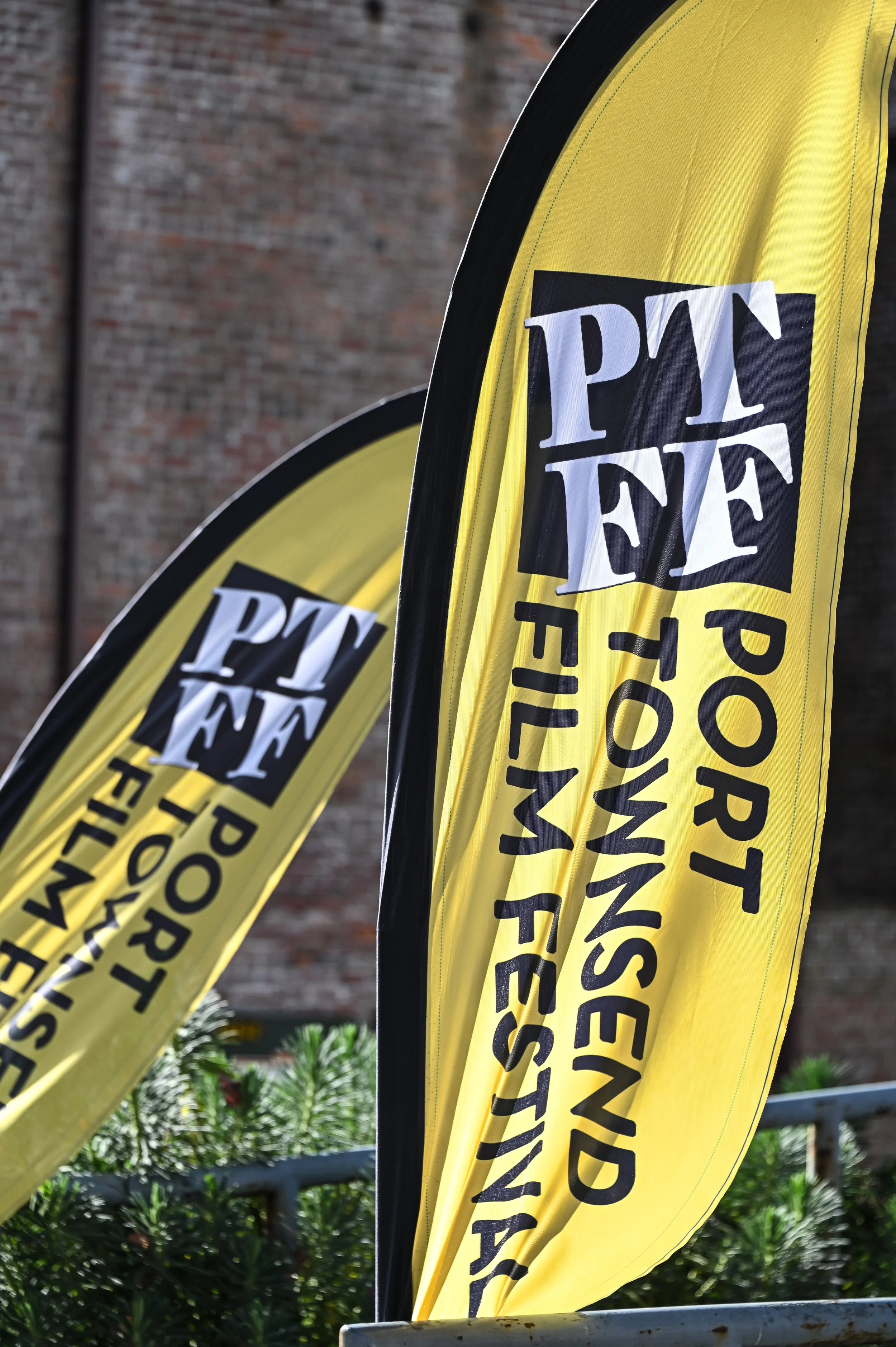 Two yellow flags with black and white text, placed outdoors near plants, against a brick wall background.