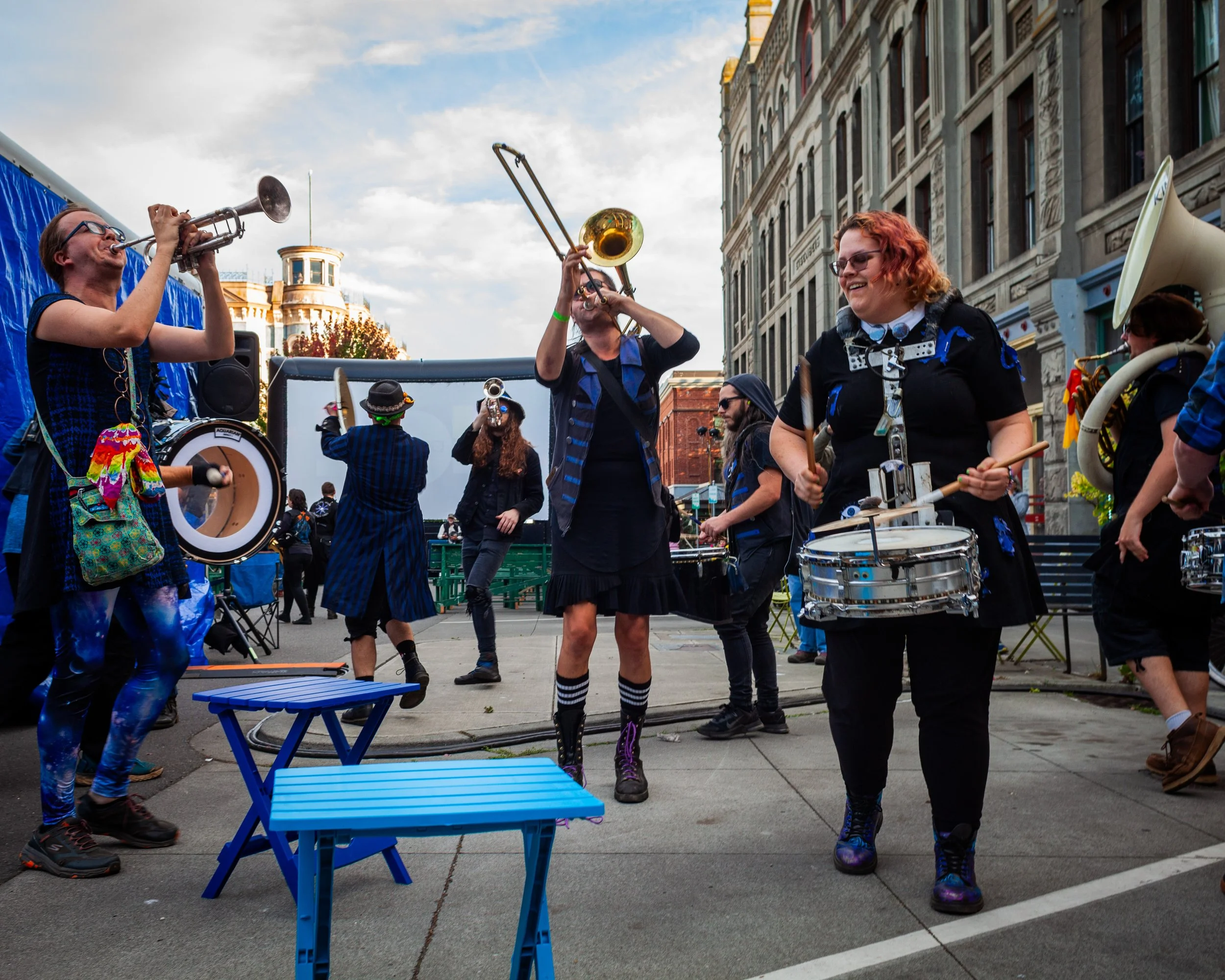 A group of street performers playing musical instruments on a city street with tall buildings in the background. They are dressed in colorful, eccentric clothing and are smiling and engaging with each other.