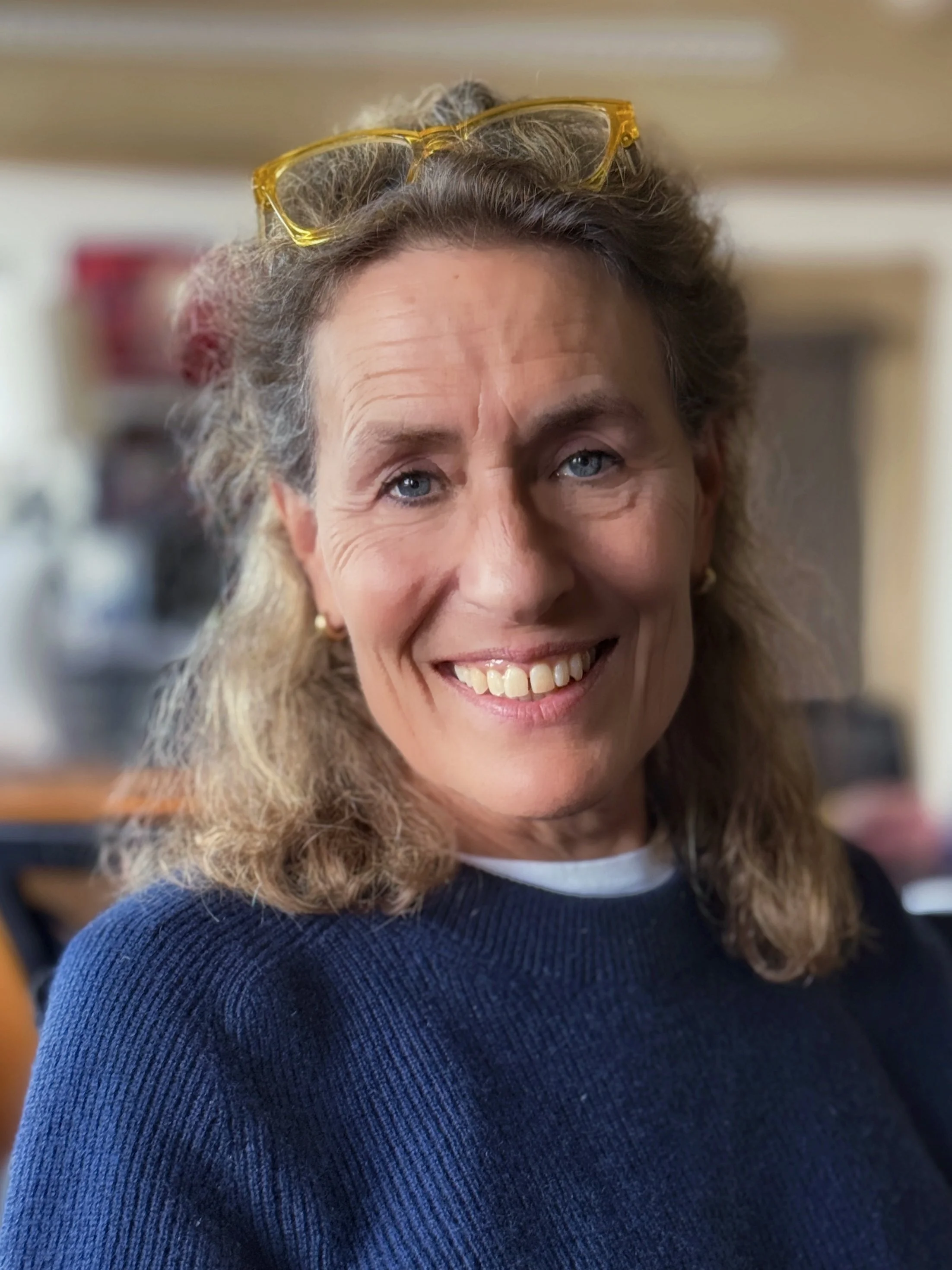 Close-up portrait of a middle-aged woman with blonde hair and blue eyes, wearing a cozy beige knit sweater, indoors.