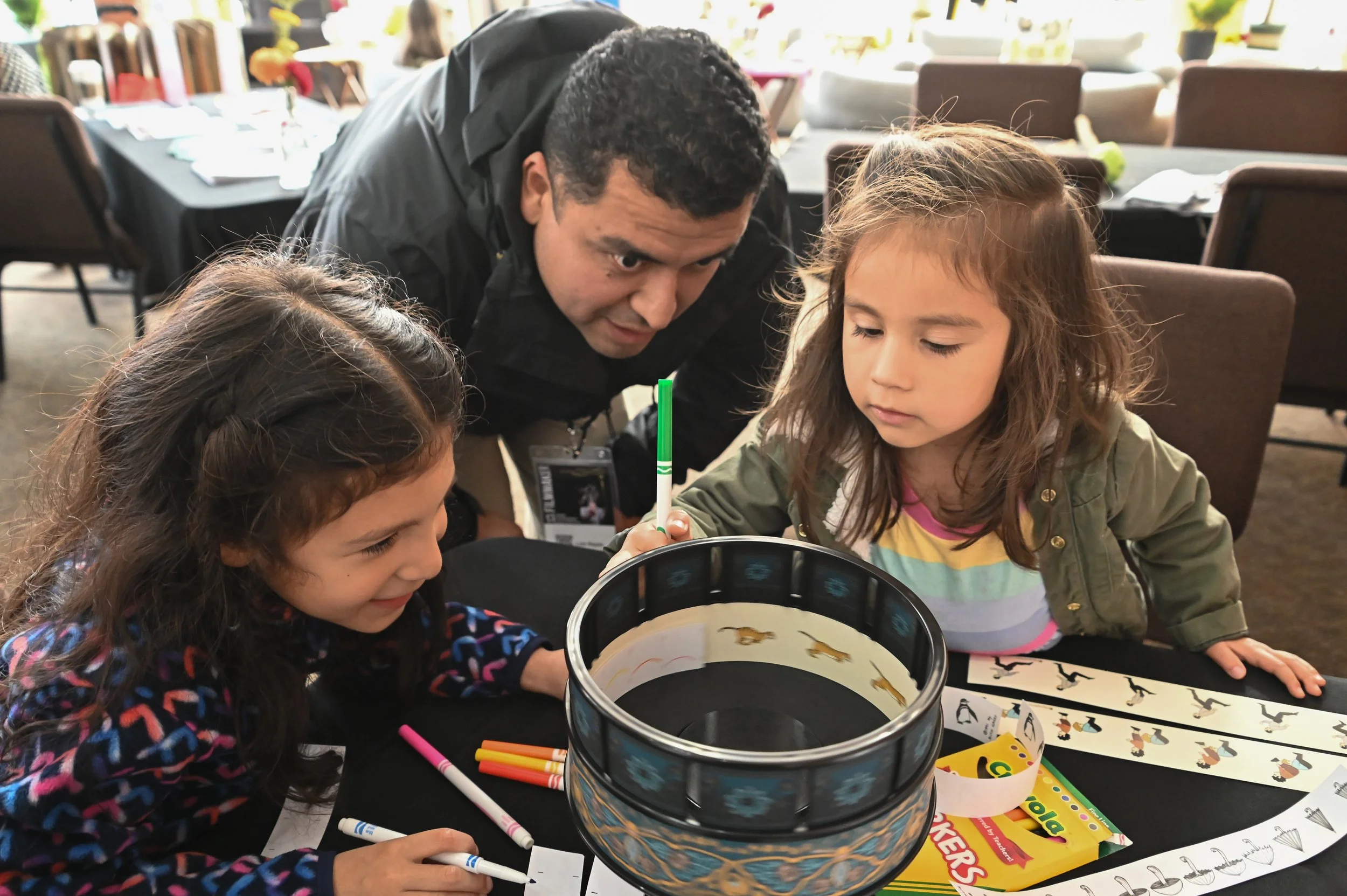 A man and two young girls sitting at a table, engaged in arts and crafts activities with markers, paper, and a bucket, in a brightly lit indoor setting with chairs and tables in the background.