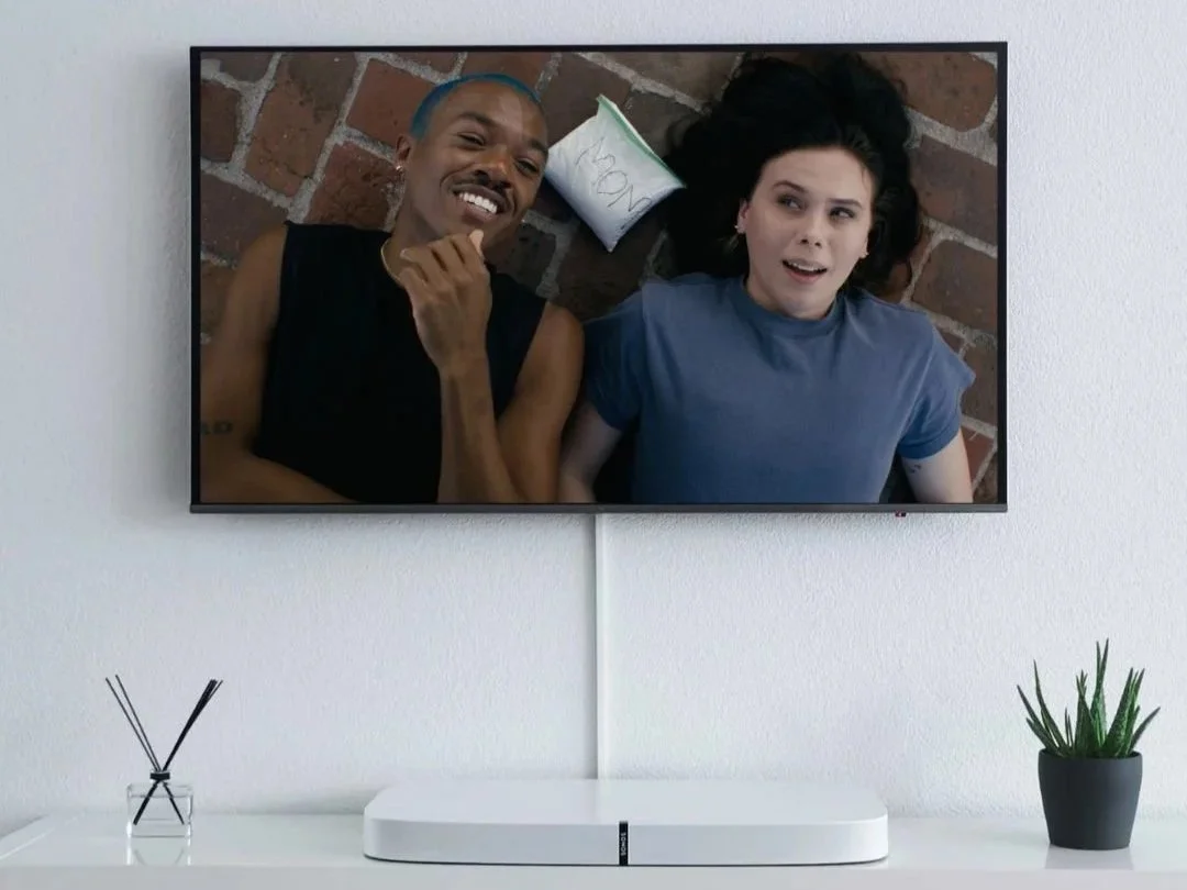 A flat-screen TV displaying a man and a woman lying on a wooden floor, smiling and looking up at the camera. There is a paper towel behind the woman and a small potted plant on a white surface below the TV.