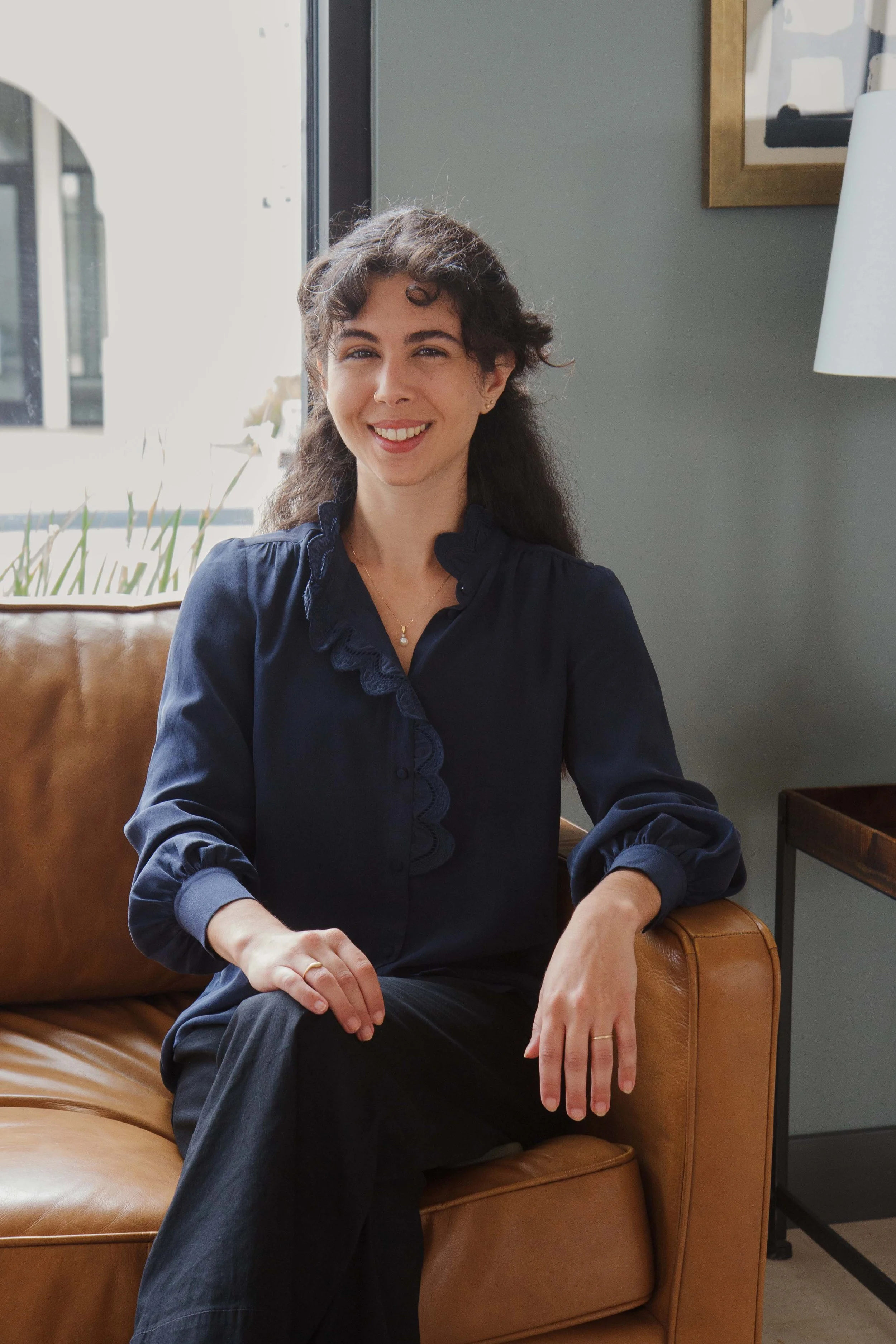 A woman with dark, slightly curly hair wearing a navy blue blouse, sitting on a tan leather sofa near a window, smiling and looking at the camera.