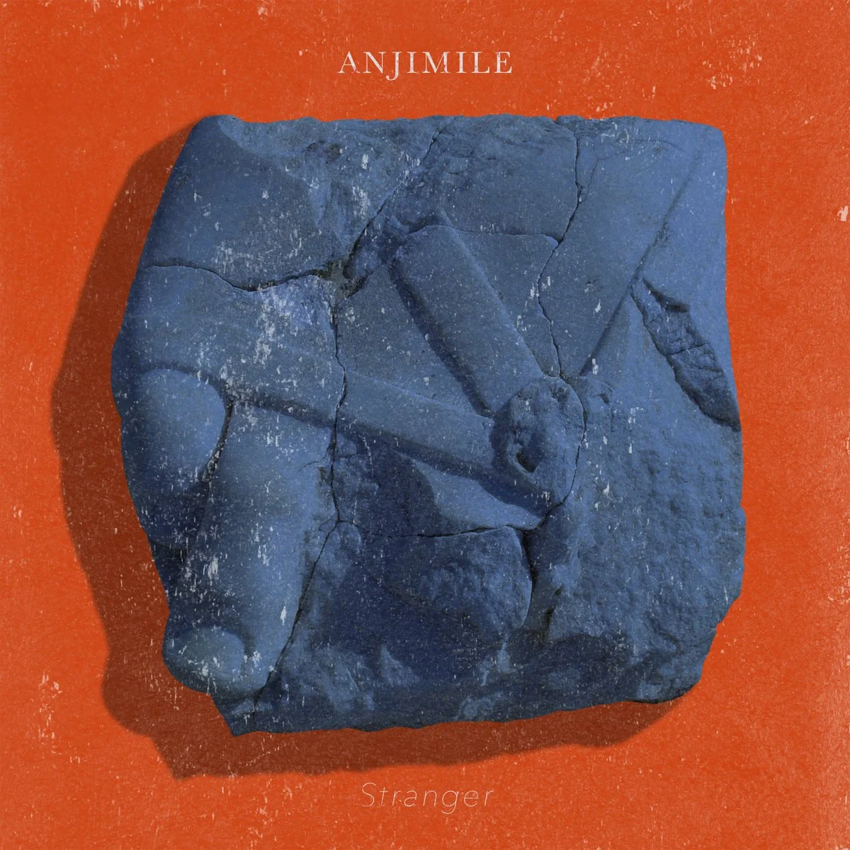 A blue stone with carved shapes on an orange background with the text 'ANJIMILE' at the top and 'Stranger' at the bottom.