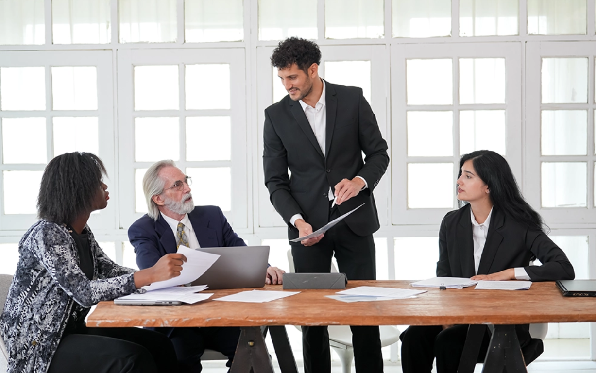 Four professionally dressed people in a meeting room with large windows. One man in a suit is standing and handing documents to a seated man with white hair and glasses. Two women, one with short curly hair and the other with long black hair, are seated at the table with papers and a laptop in front of them.