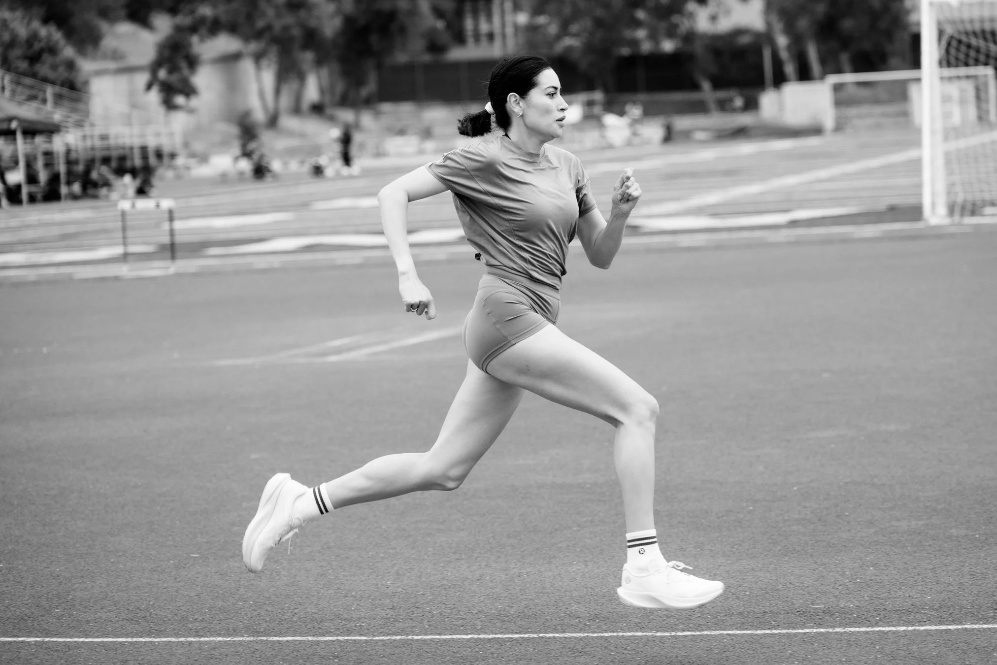A woman running on an outdoor track, wearing athletic clothes and sneakers, with trees and a fence in the background.