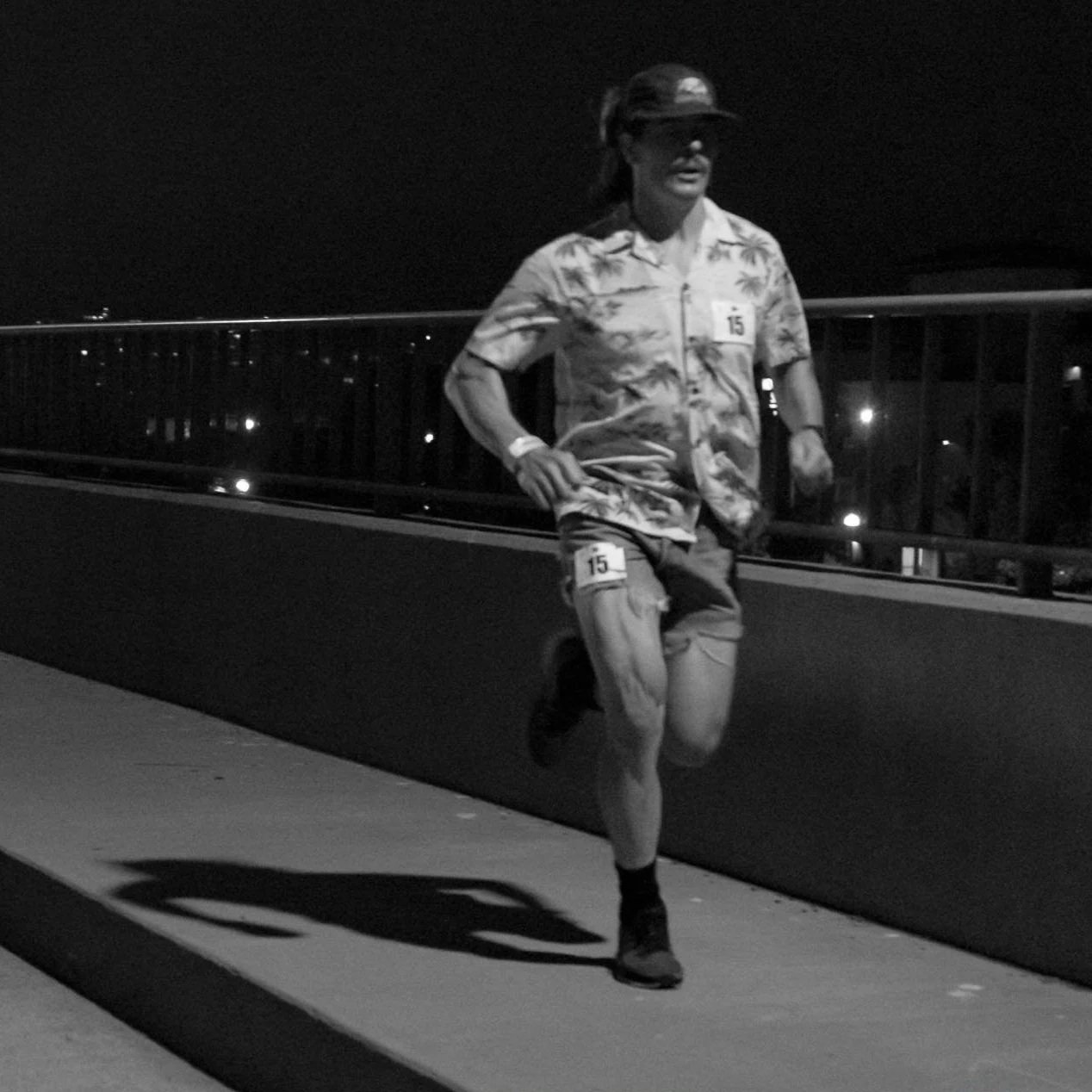 A person running at night on a bridge, wearing sunglasses, a Hawaiian shirt, shorts, and a hat, with race bibs numbered 15 on their shirt and shorts.