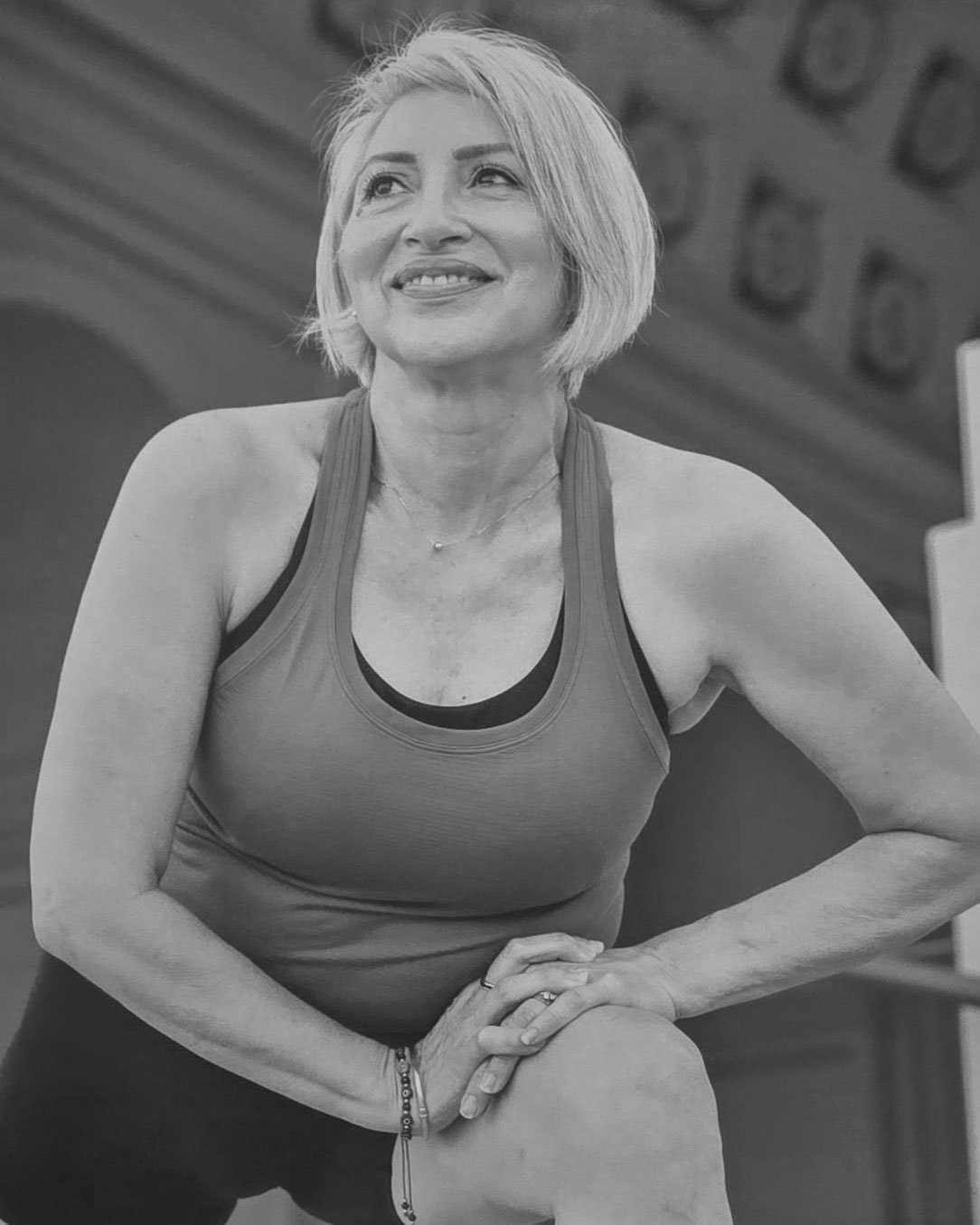 A smiling middle-aged woman with short blonde hair in workout attire, resting her hands on her knee, in a gym setting.