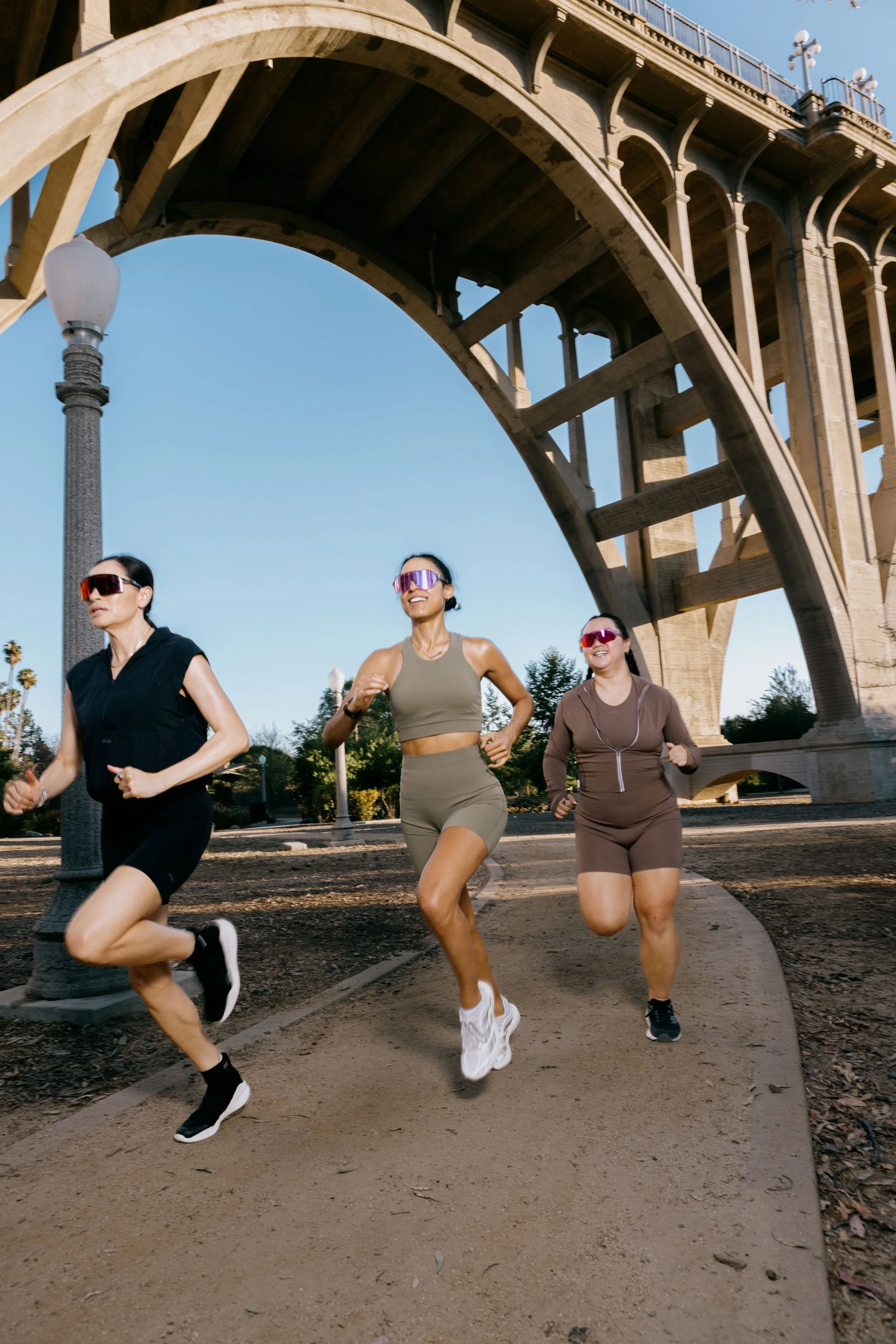 Three women jogging together outdoors in a park area near a large concrete bridge structure, with trees and lamp posts in the background.