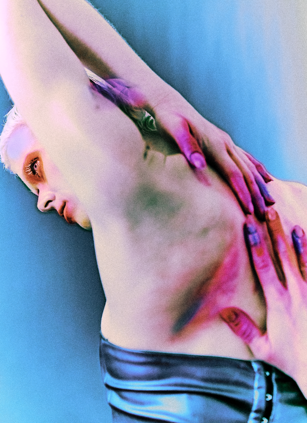A close-up image of a person's torso with colorful lighting effects, featuring their hand placed on their chest and shoulder, against a bluish background.