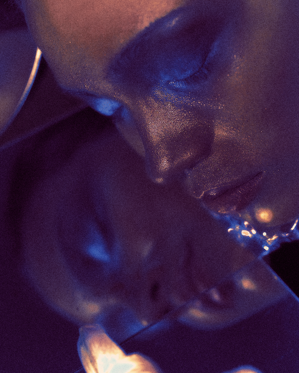 Close-up of a person's face with glitter, with their eyes closed and head tilted downward, in low, colorful lighting.