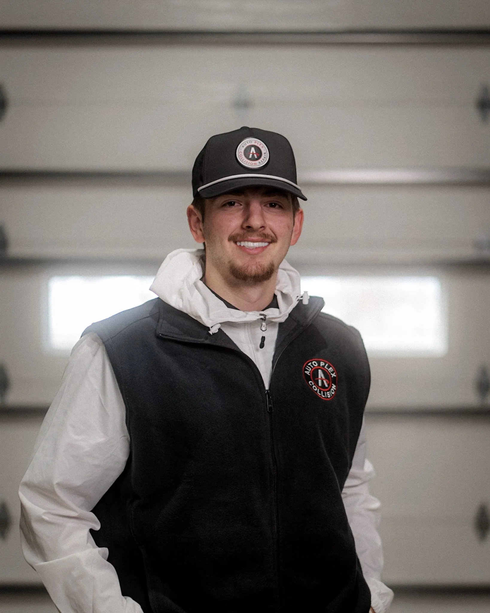 A young man standing in front of a garage door, wearing a black Columbia vest with a logo that says Auto Plex and a black cap with a round emblem. He has a smile and his hands in his pockets.