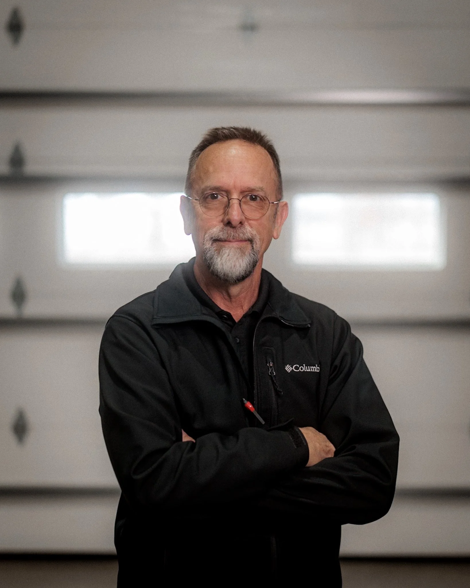 A man with glasses and a beard wearing a black cap and black athletic jacket with a logo, standing with arms crossed in a garage with a garage door in the background.