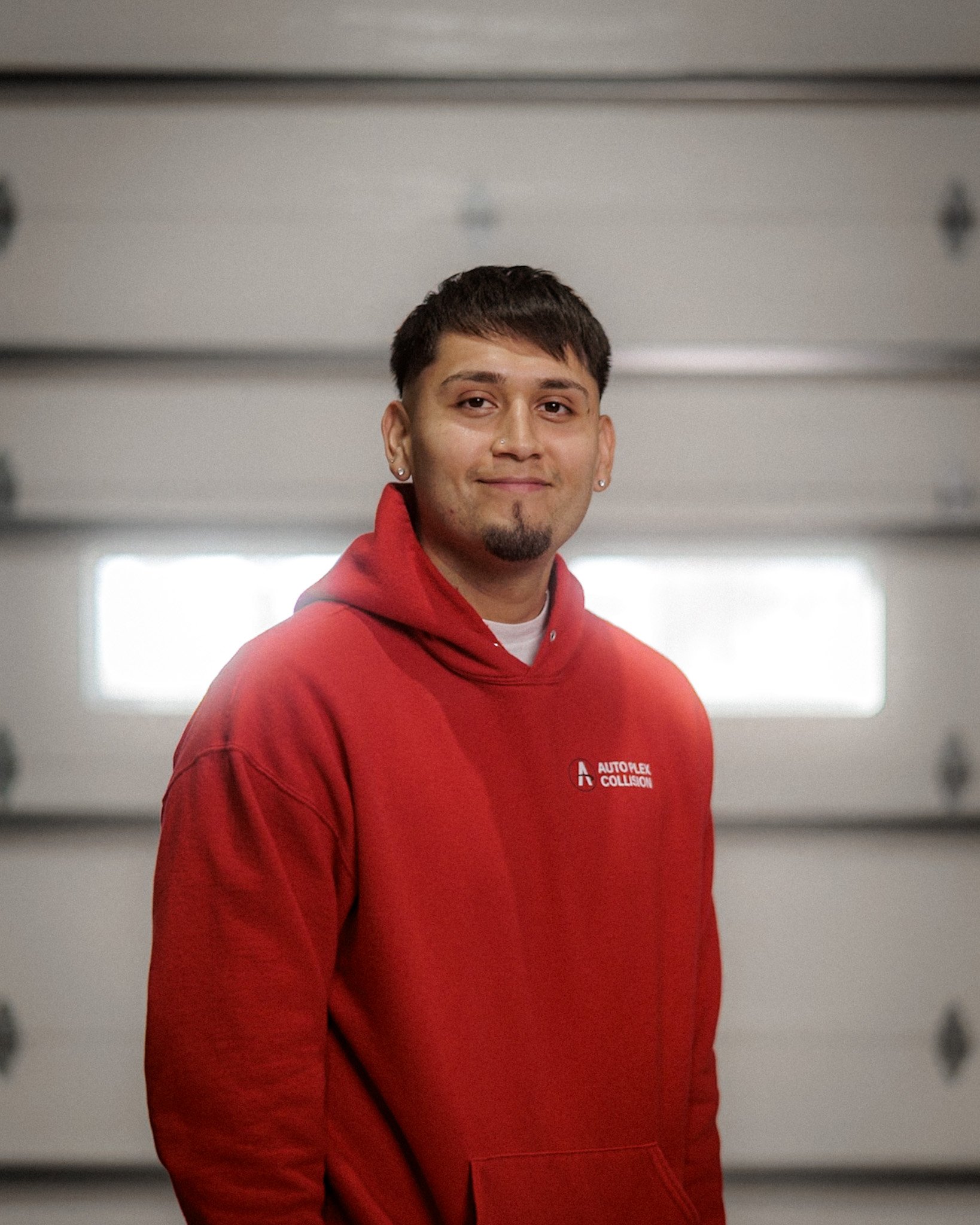 A young man with dark hair, a goatee, and light skin standing inside a garage with a closed door in the background, wearing a gray and black Columbia jacket and smiling at the camera.
