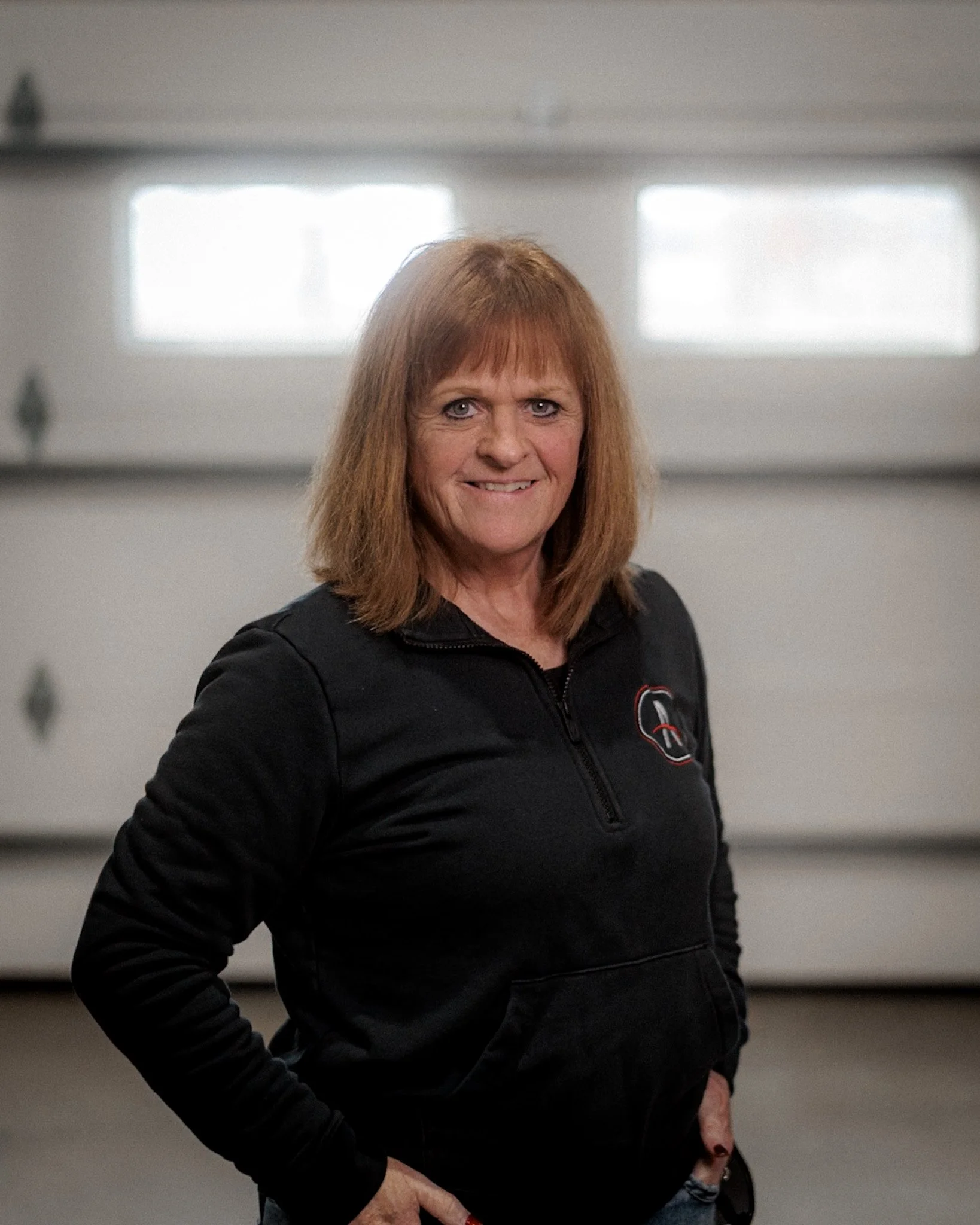 A woman with shoulder-length brown hair standing inside a garage, wearing a dark gray polo shirt with a logo on the chest.