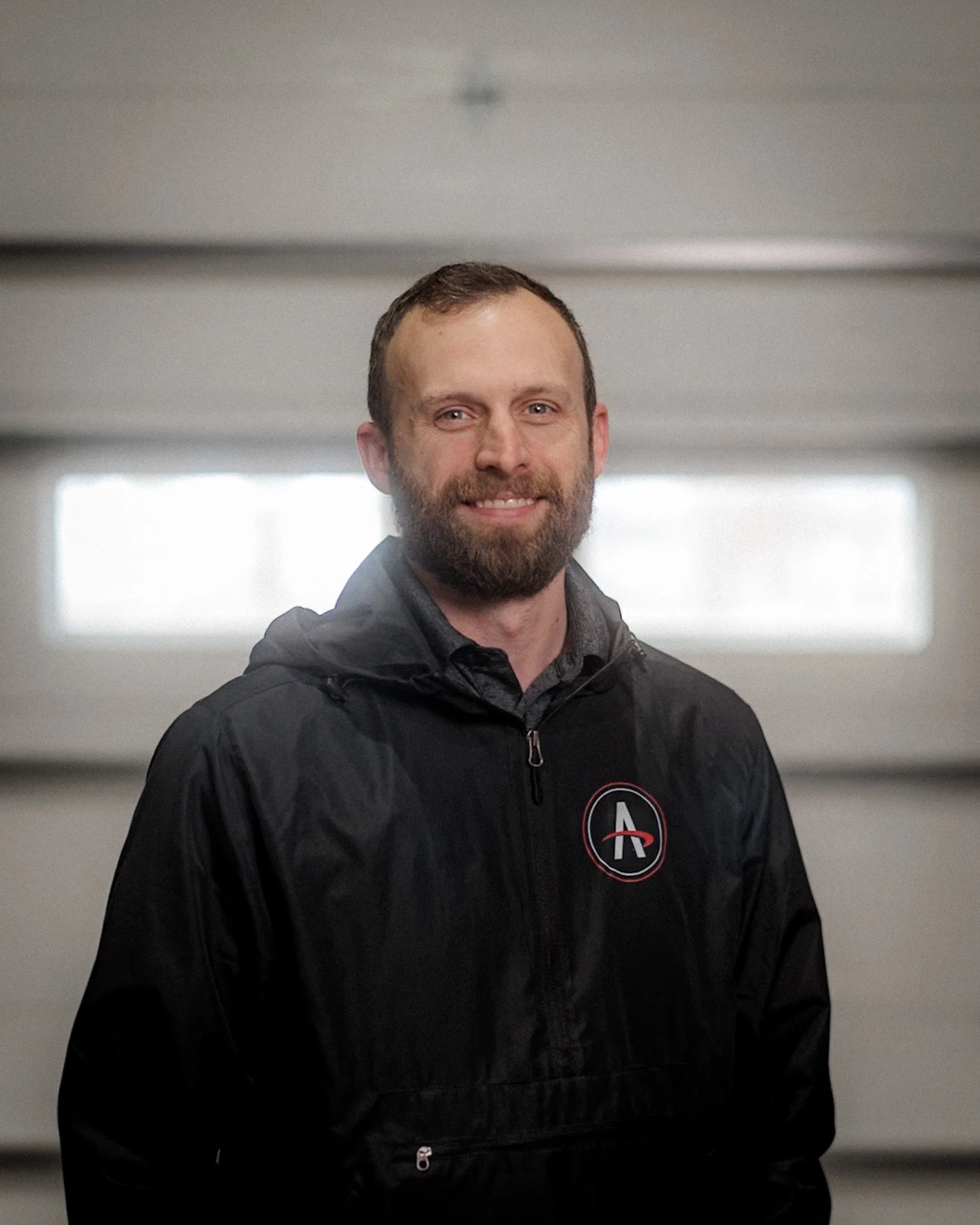 Smiling man with a beard and short hair, wearing a black athletic jacket, standing inside a garage with a white roll-up door in the background.