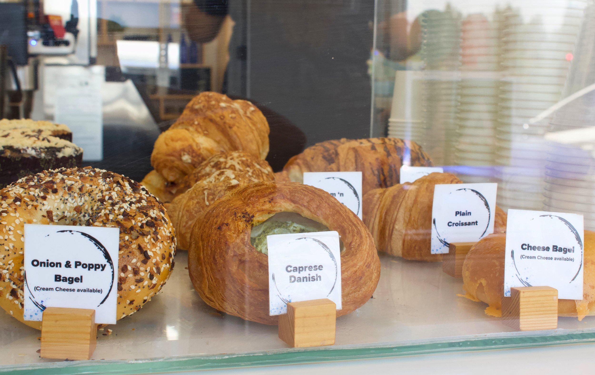 pastries, including a bagel, a caprese danish, and a croissant in a pastry box in coffee shop behind glass.