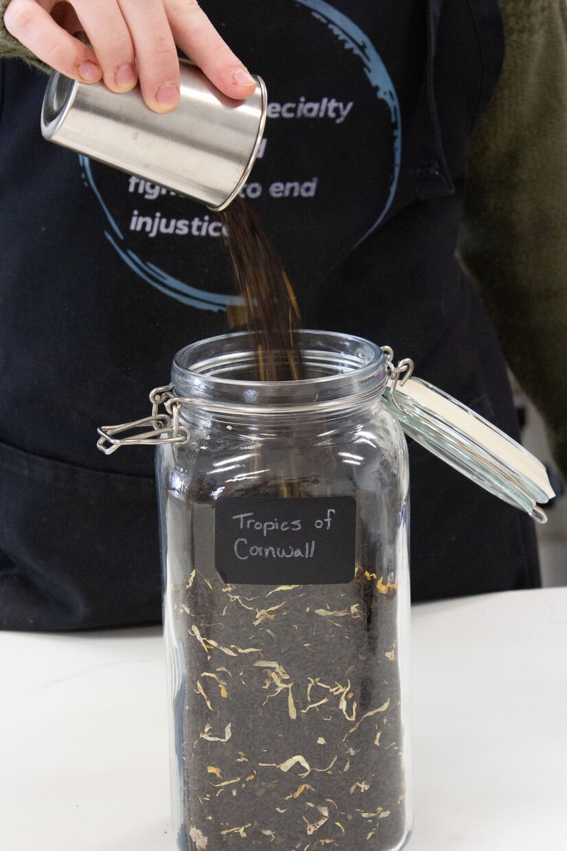 pouring in loose leaf tea called "tropics of cornwall" into a glass jar with label on it. tea has papaya, pineapple, and black tea in it.
