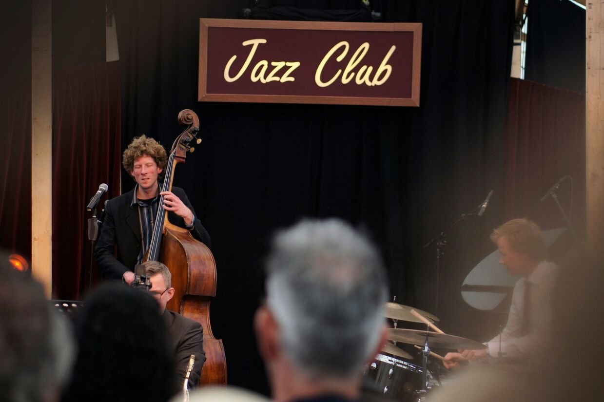 A man playing a chello with a drummer and a "Jazz Club" sign in the background.