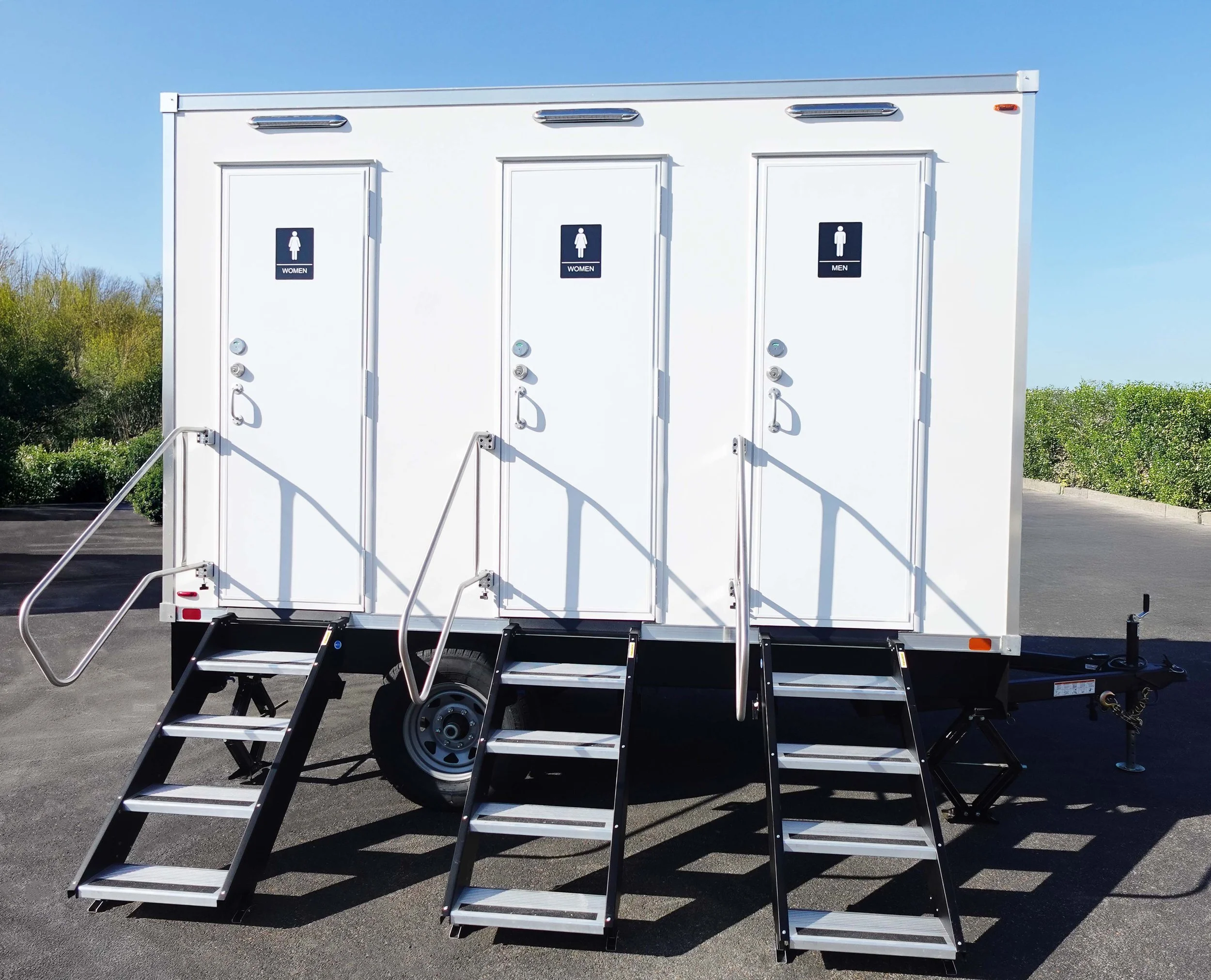 Portable restroom facility with separate entrances for women and men, accessible stairs, and handrails, situated on a paved surface outdoors with green bushes and a blue sky in the background.