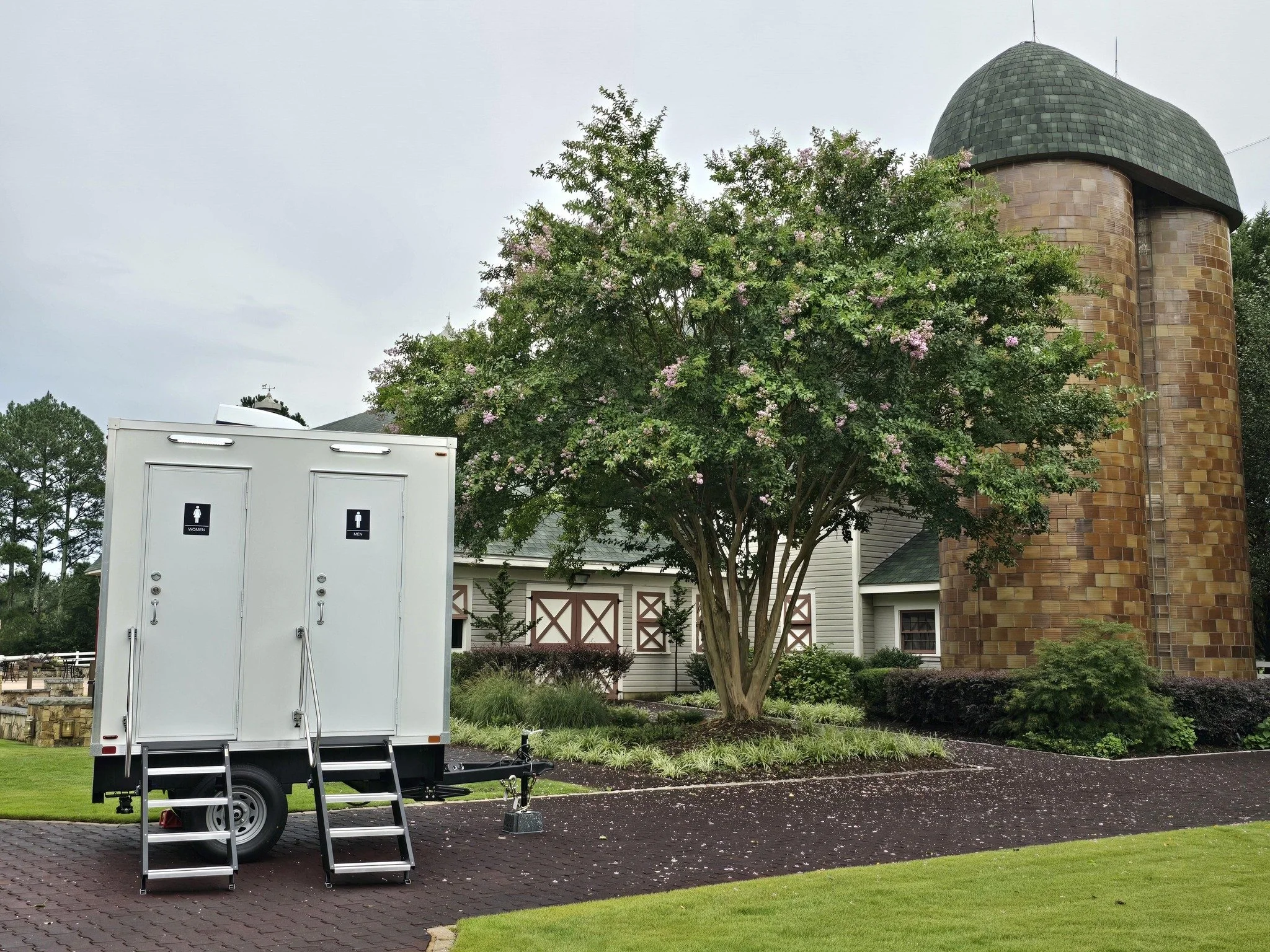 Portable restroom trailers with stairs in front, garden with trees and bushes, white house with shutters, and a large rounded brick building in the background.