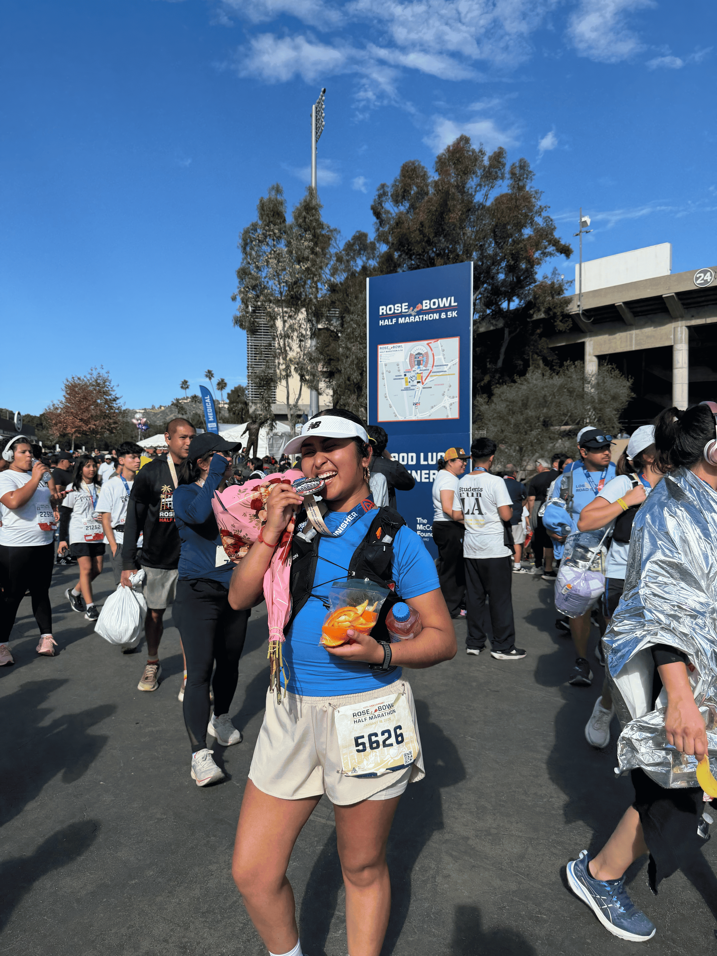 A young woman with a race bib eating a medal at the Rose Bowl Half Marathon & 5K event, surrounded by other runners and spectators outdoors on a sunny day with a blue sky.