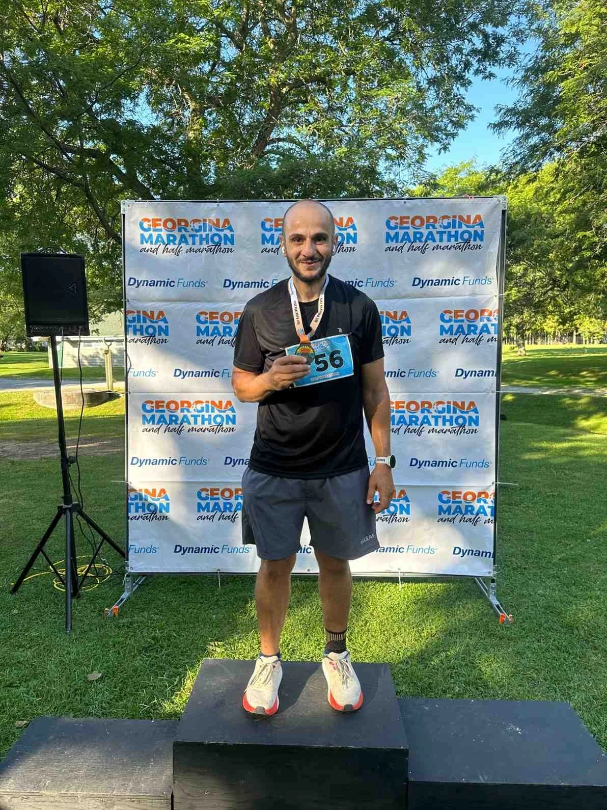 Man standing on a podium holding a medal after completing the Georgia Marathon, posing in front of a backdrop with event logos, in a park with green grass and trees.