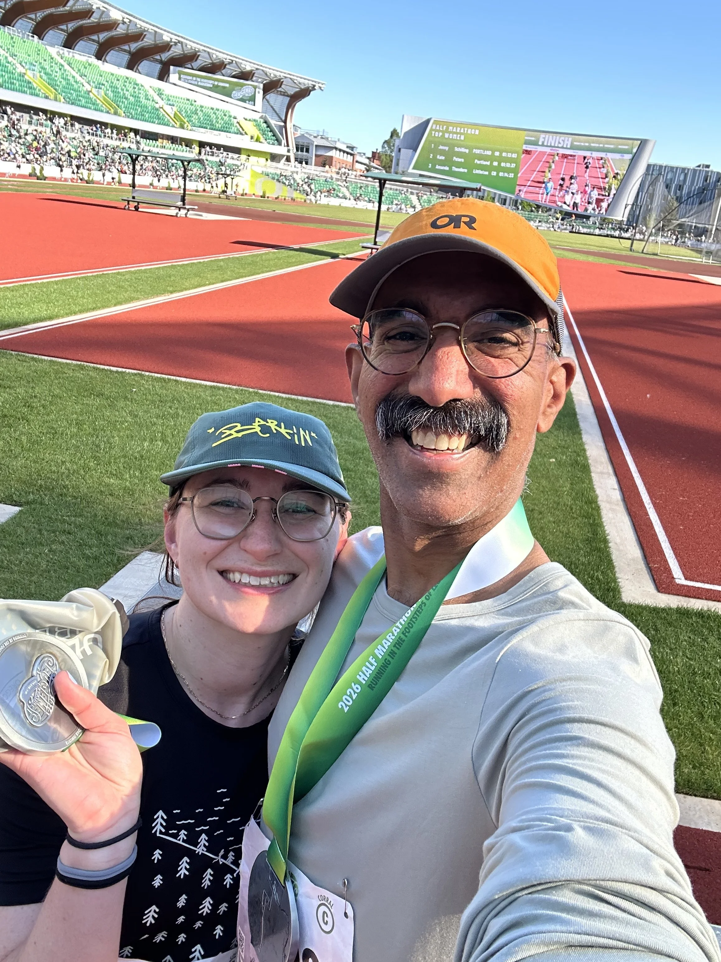 A man and a woman taking a selfie at a track and field stadium after completing a race. The woman is holding a medal. The man is wearing a cap and glasses, and the woman has freckles and wears glasses. In the background, there are green stadium seats and a large electronic scoreboard displaying race results, with a bright blue sky above.