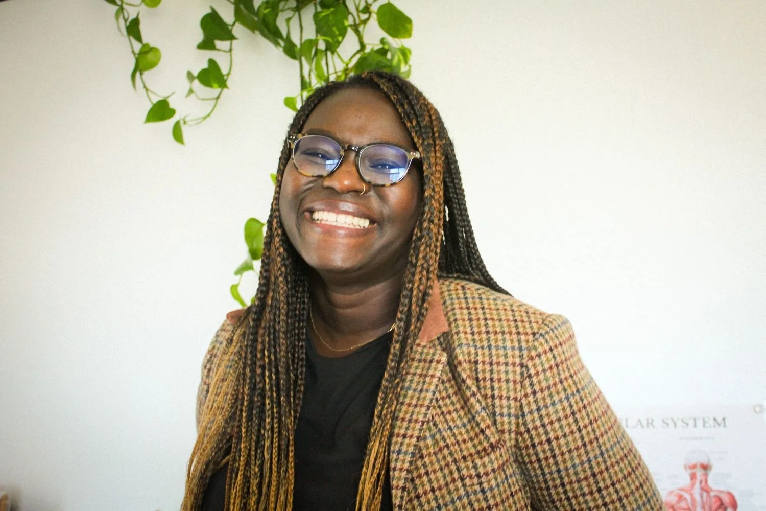 A smiling Black woman with braided hair, wearing glasses, a black shirt, and a brown checked blazer, standing indoors in front of a white wall with green hanging plants.