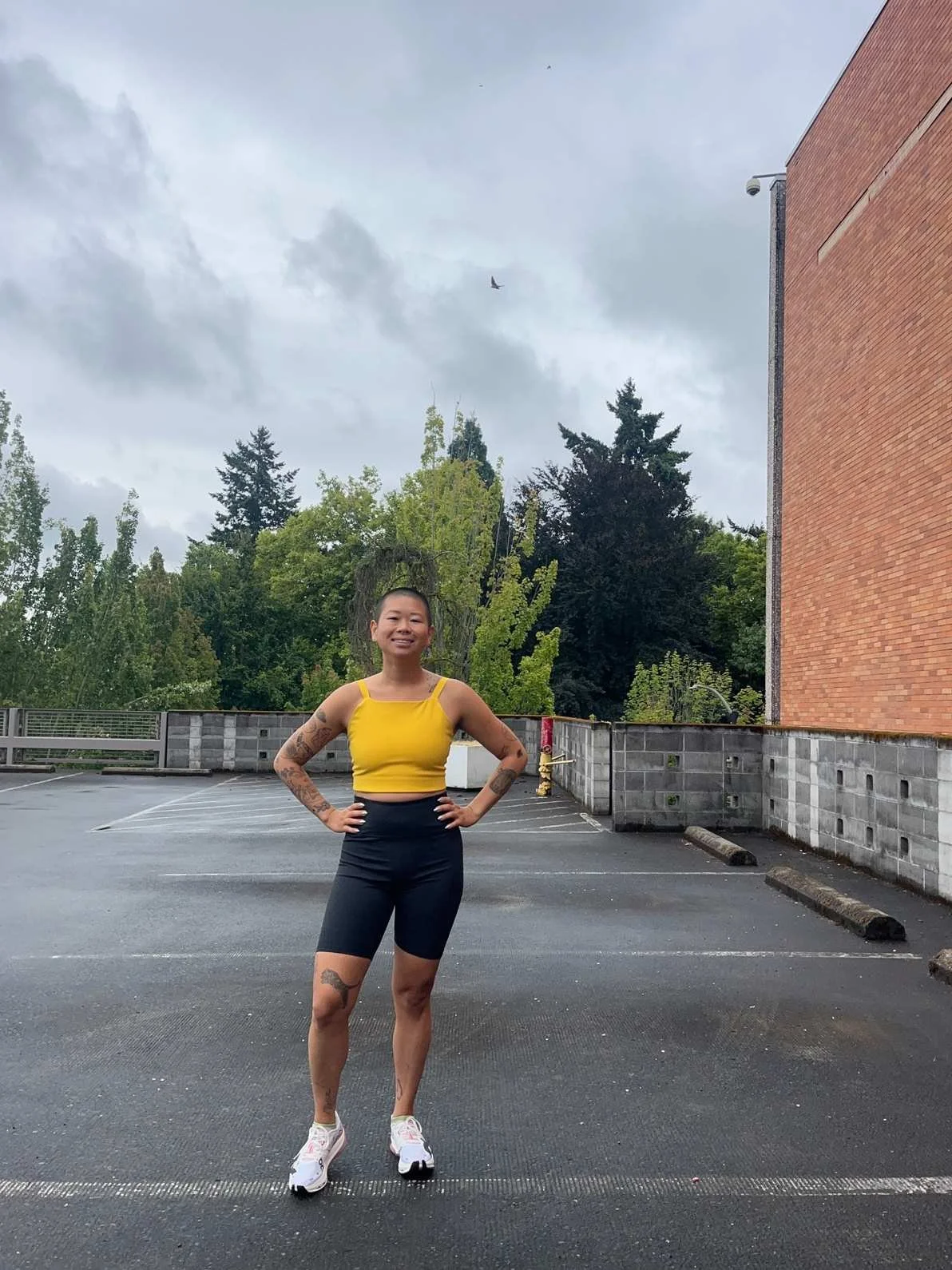 A woman with tattoos in a yellow crop top and black shorts standing with hands on hips in an empty parking lot outdoors on a cloudy day with trees and a brick building in the background.