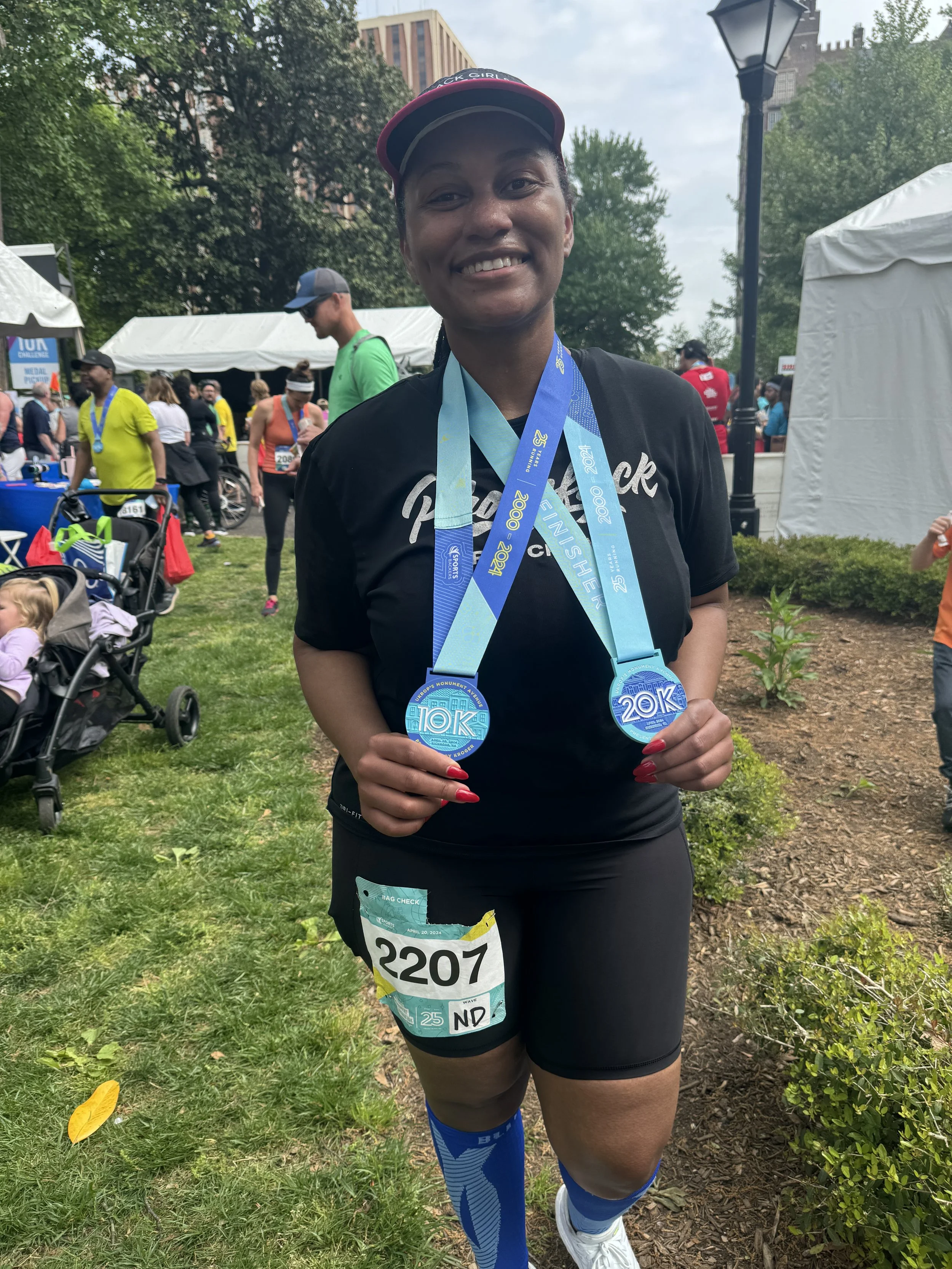 A woman smiling and holding two race medals, standing outdoors at a race event, wearing black athletic clothes, race bib number 2207, with other runners and event tents in the background.