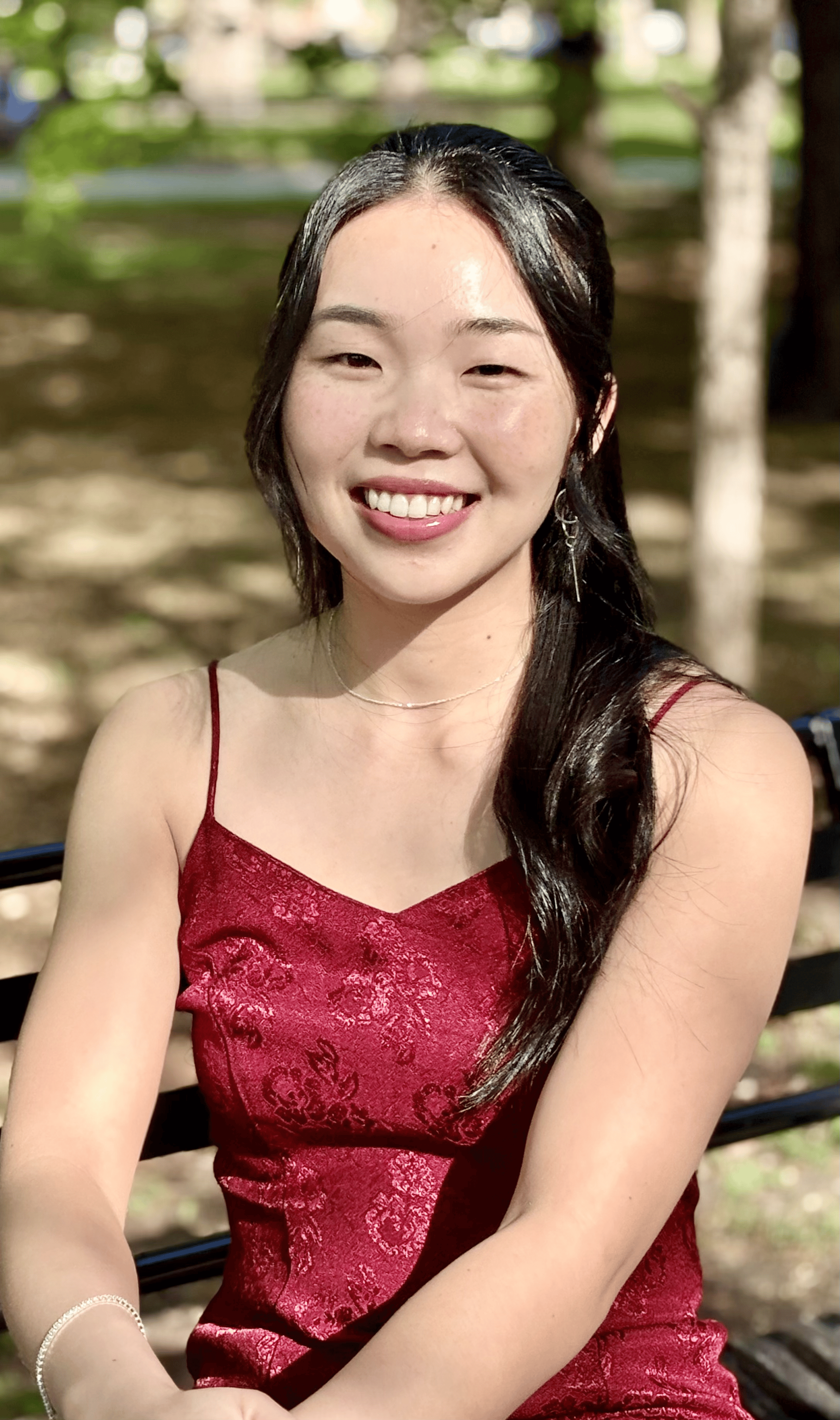 A young woman with long dark hair wearing a red dress with spaghetti straps, sitting on a bench outdoors with trees and sunlight in the background, smiling at the camera.