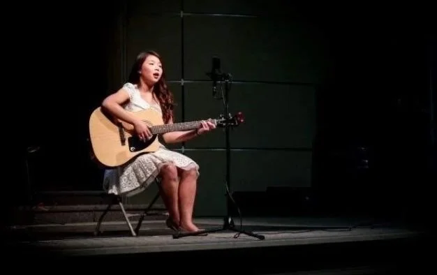 A young girl sitting on a chair playing an acoustic guitar and singing into a microphone on a stage with a dark background.