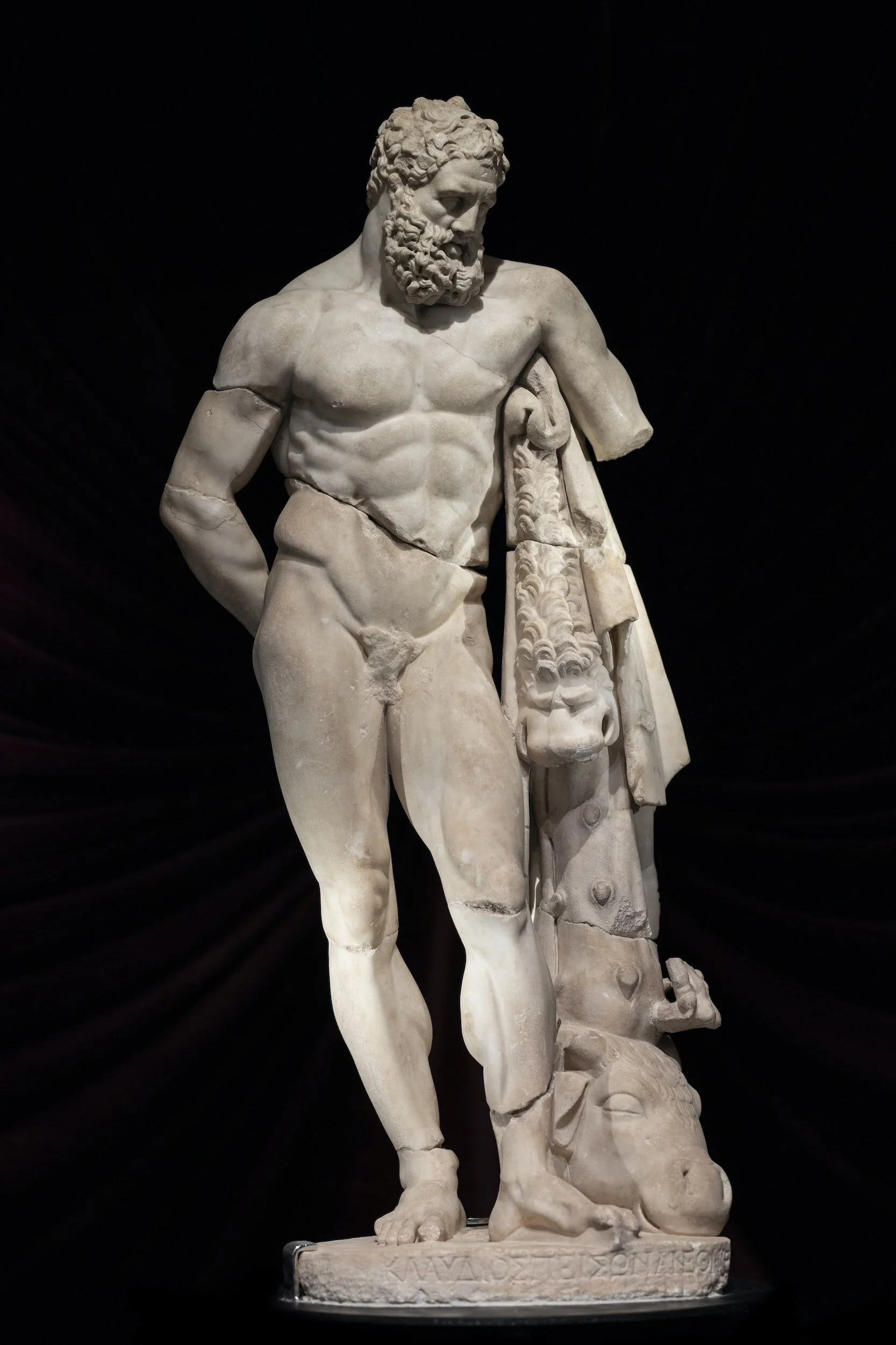 Ancient Greek marble sculpture of a muscular, bearded man holding a draped garment with a lion's head and other decorative elements, standing on a base.