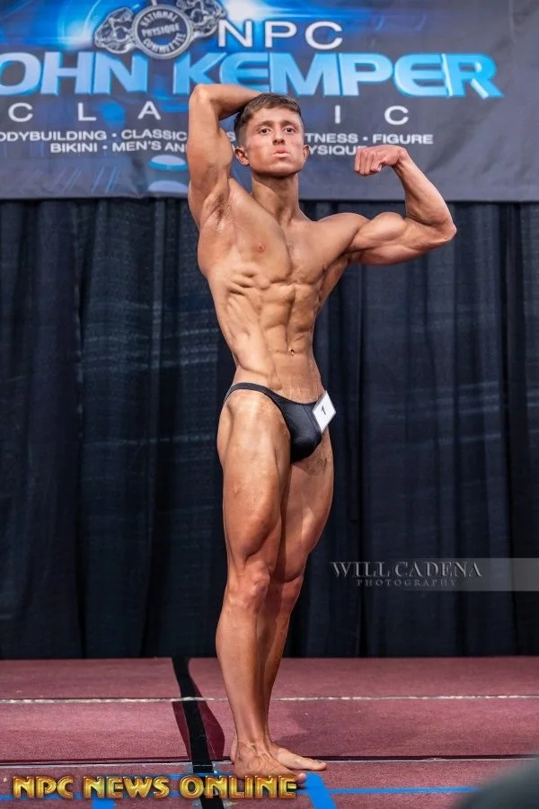 Male bodybuilder on stage in black posing trunks, striking a side chest pose, with visible muscles and defined abs, against a backdrop with NPC and John Kemper logos.