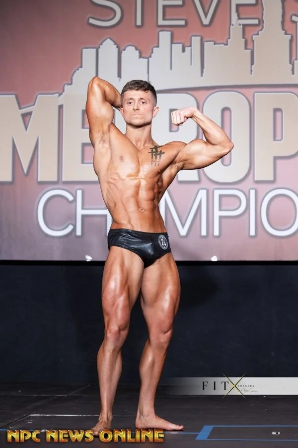 A male bodybuilder posing on stage at a bodybuilding competition, wearing black shorts with the number 24, in front of a backdrop with the words "Mr. Olympia" and cityscape graphics.