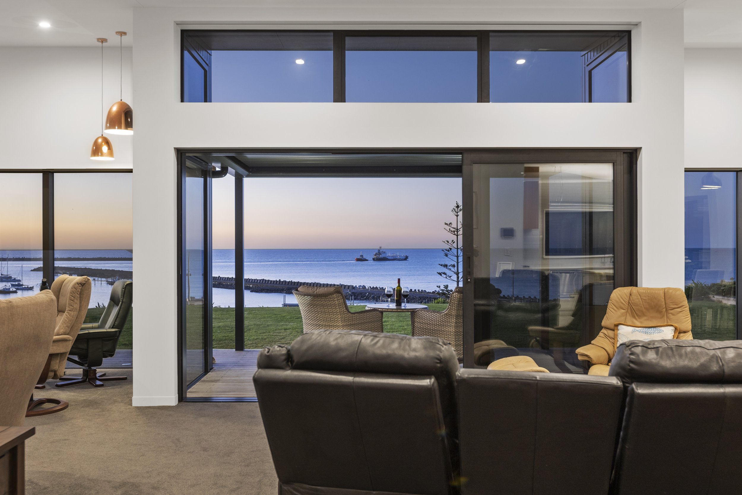 Modern living room interior with large windows overlooking an ocean view at sunset. The room features leather and fabric chairs, pendant lights, and a small outdoor seating area.