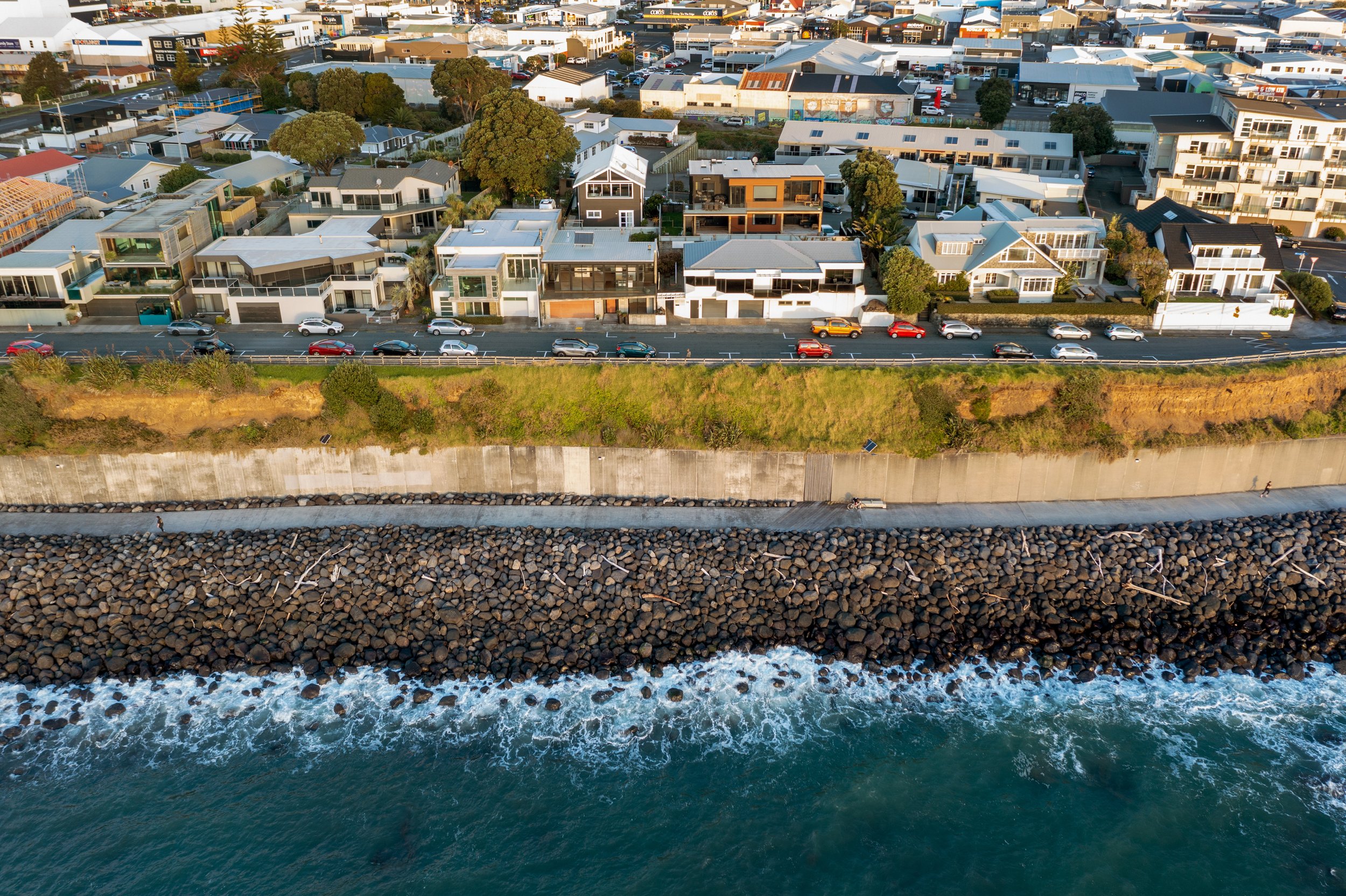 Aerial view of a coastal neighborhood featuring modern houses, a road with cars, a grassy cliff, and a rocky shoreline next to the ocean.