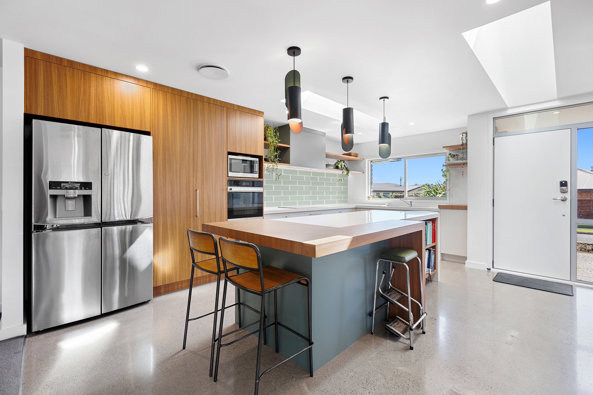 Modern kitchen with stainless steel refrigerator, wooden cabinets, island with bar stools, pendant lights, and green tile backsplash.
