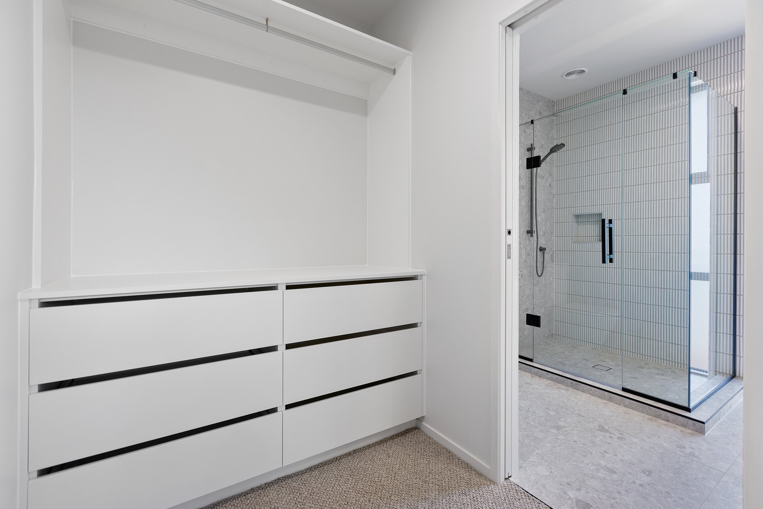 Minimalist white wardrobe with multiple drawers next to a modern bathroom featuring a glass-enclosed shower and tiled walls.