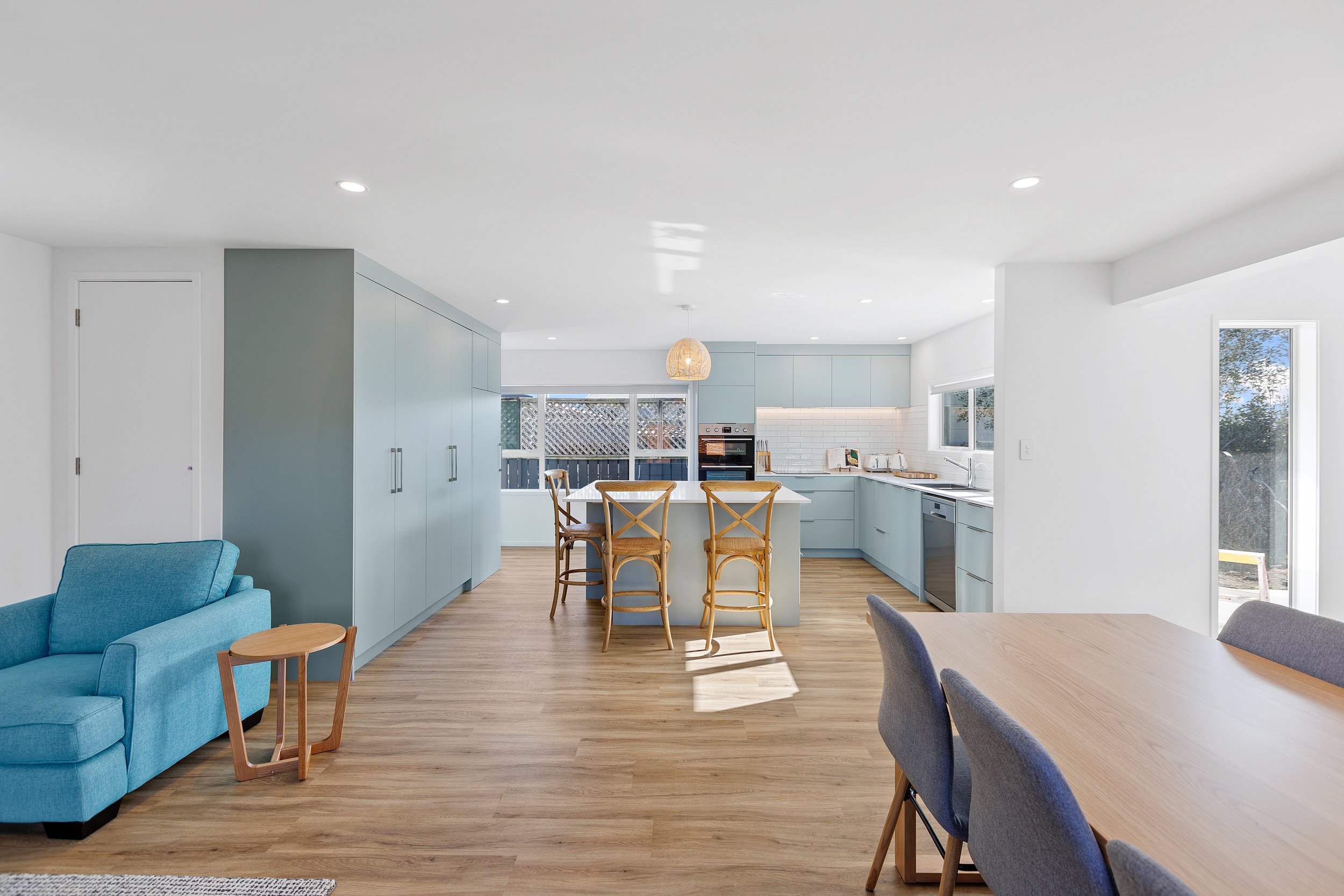 Modern open-plan kitchen and dining area with blue cabinets, wooden flooring, and a light blue armchair; features an island with wicker chairs and a dining table with gray chairs.