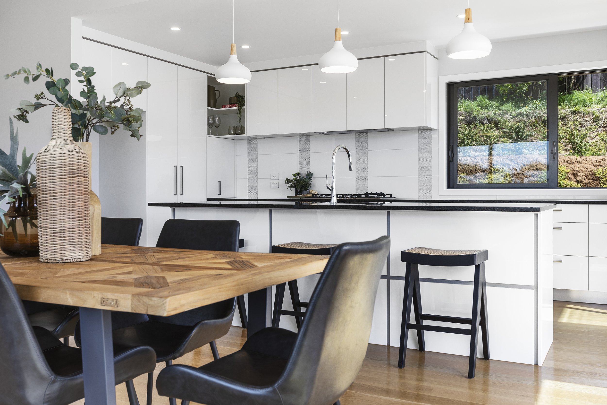 Modern kitchen and dining area with white cabinets, black countertops, wooden dining table, black chairs, pendant lighting, and a large window with greenery outside.